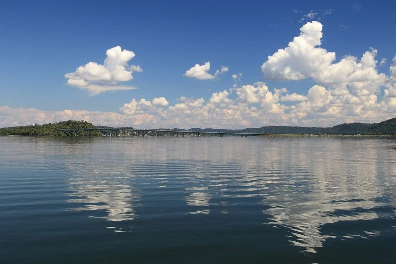 Calm lake reflecting clouds with green hills and forested shoreline under a blue sky.