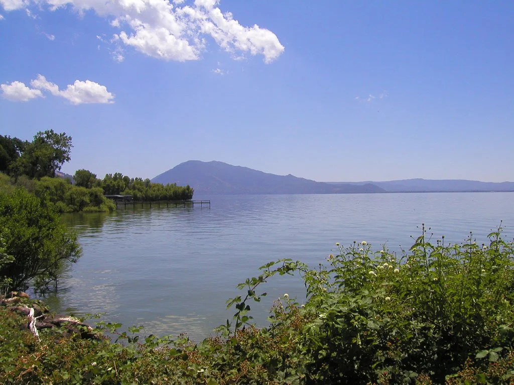 Scenic view of Clear Lake, California, with calm water, surrounding greenery, and mountainous backdrop under a blue sky with few clouds.