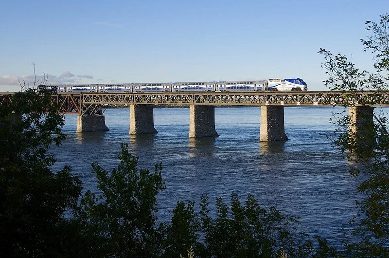 Train crossing a bridge over a river with trees in the foreground.