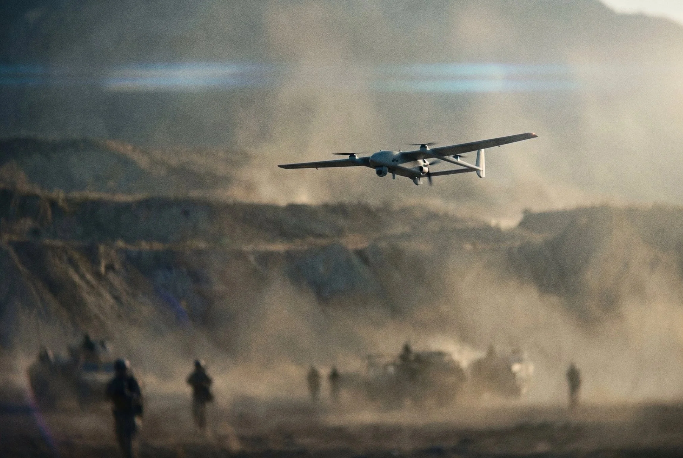 Fixed-wing drone flying low over a dusty rugged landscape with vehicles and personnel visible below.