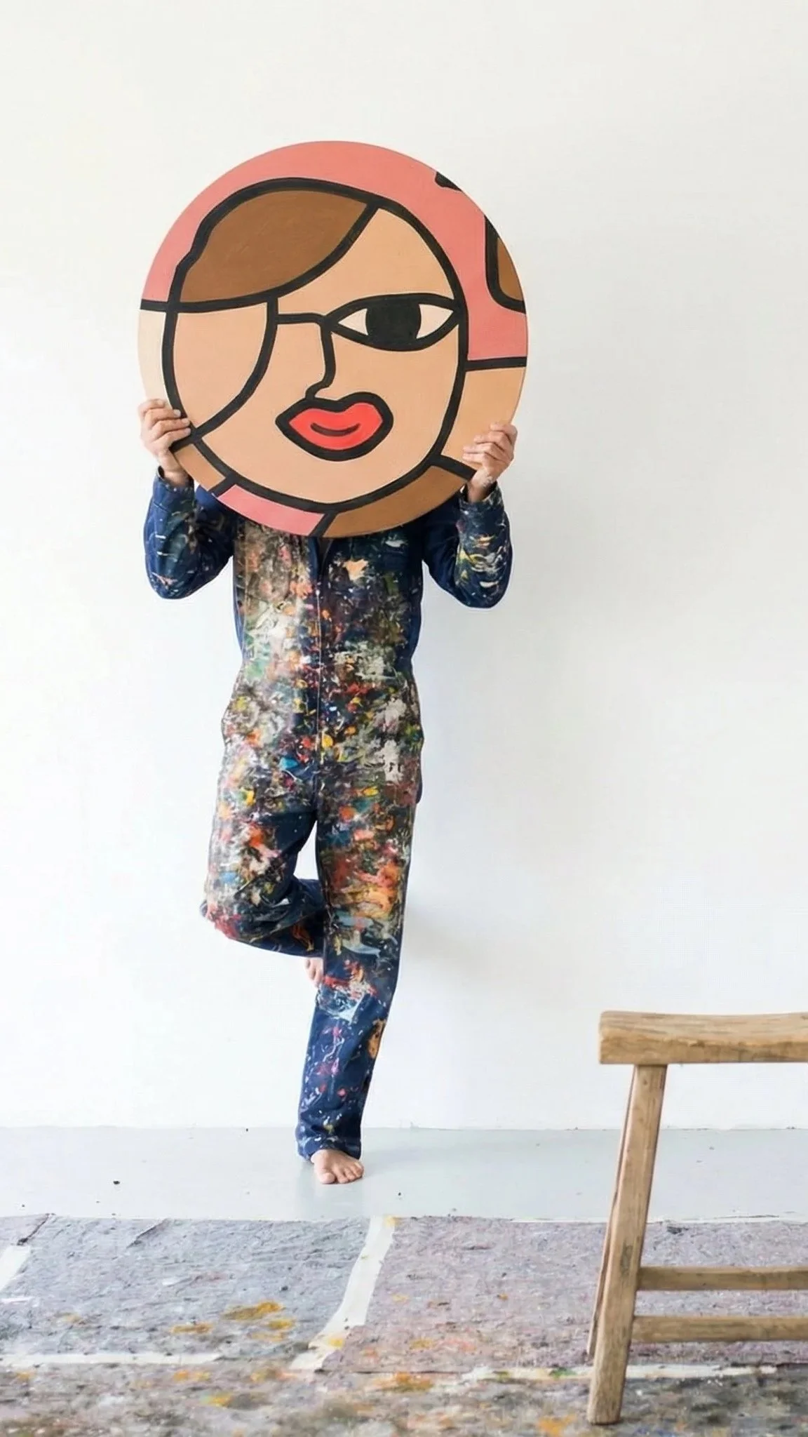 Person in paint-splattered clothes holding a large round artwork of a stylized face with one eye, glasses, and red lips in front of their face.