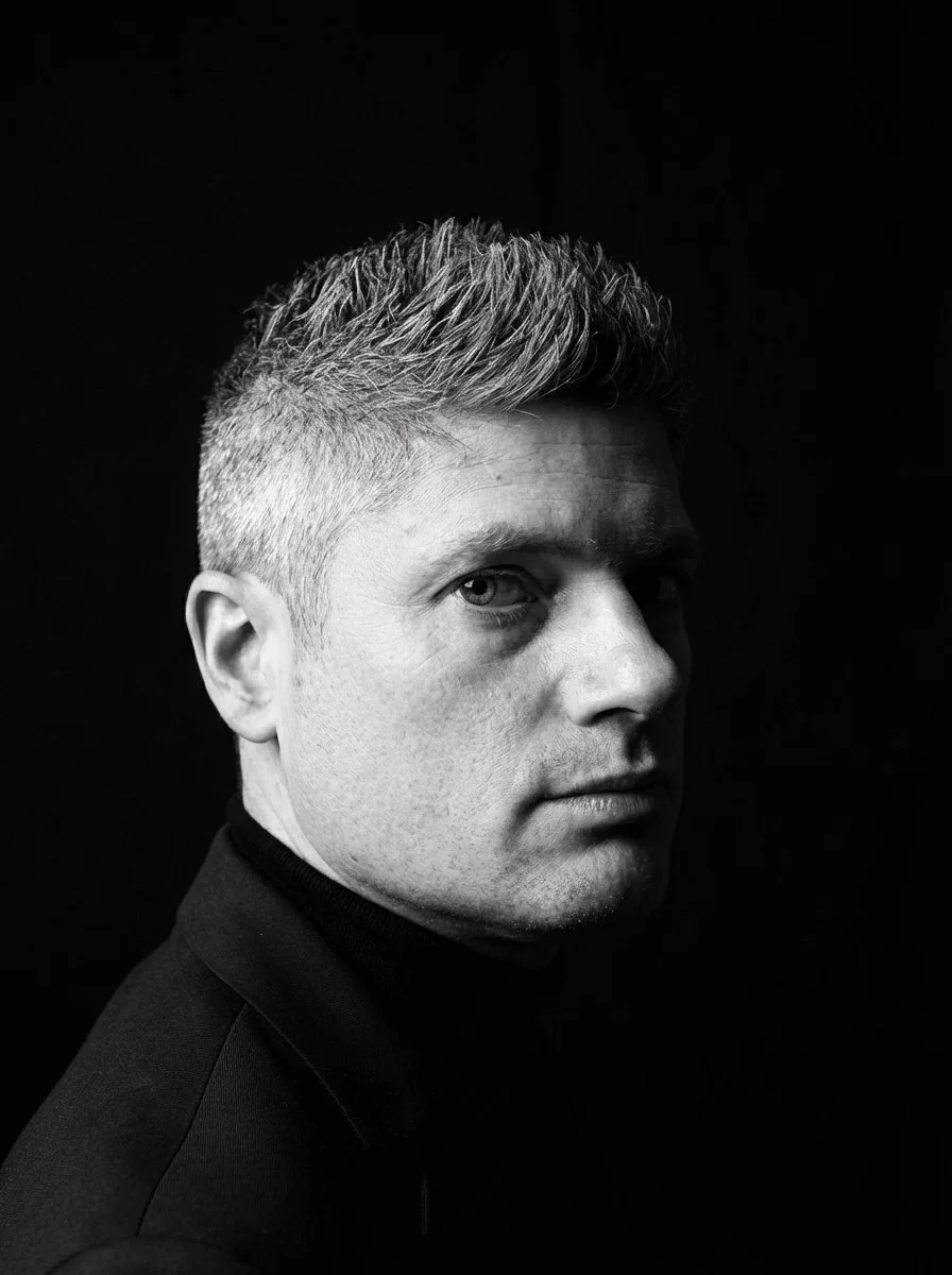 Black and white portrait of a man with short, textured hair, wearing a dark turtleneck, looking over his shoulder against a dark background.