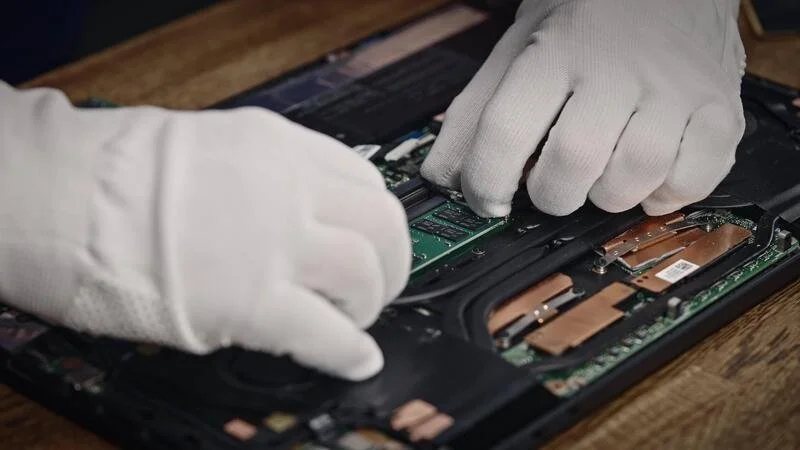 Hands wearing white gloves repairing a laptop with the interior components exposed.