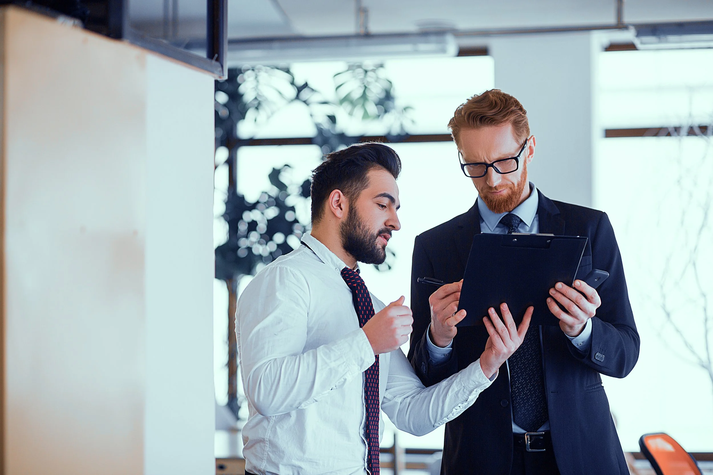 Two businessmen, one in a white shirt and the other in a dark suit, are looking at a tablet in a modern office setting.