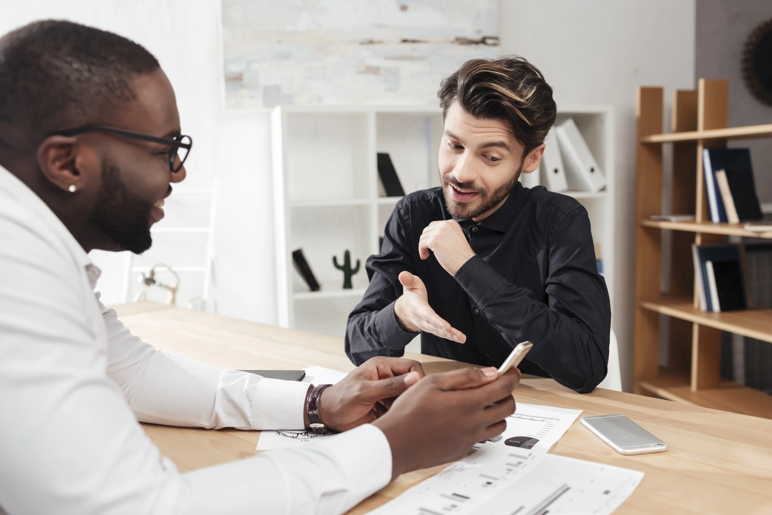 Two men sitting at a wooden table in a modern office, having a discussion while looking at a smartphone. One man is wearing glasses and a white shirt, the other wears a black shirt. Documents and two smartphones are on the table.