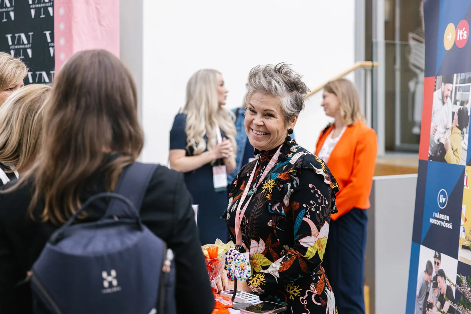 Nainen keskustelee muiden ihmisten kanssa tapahtuma-alueella, jossa on banneri ja ihmisiä taustalla.