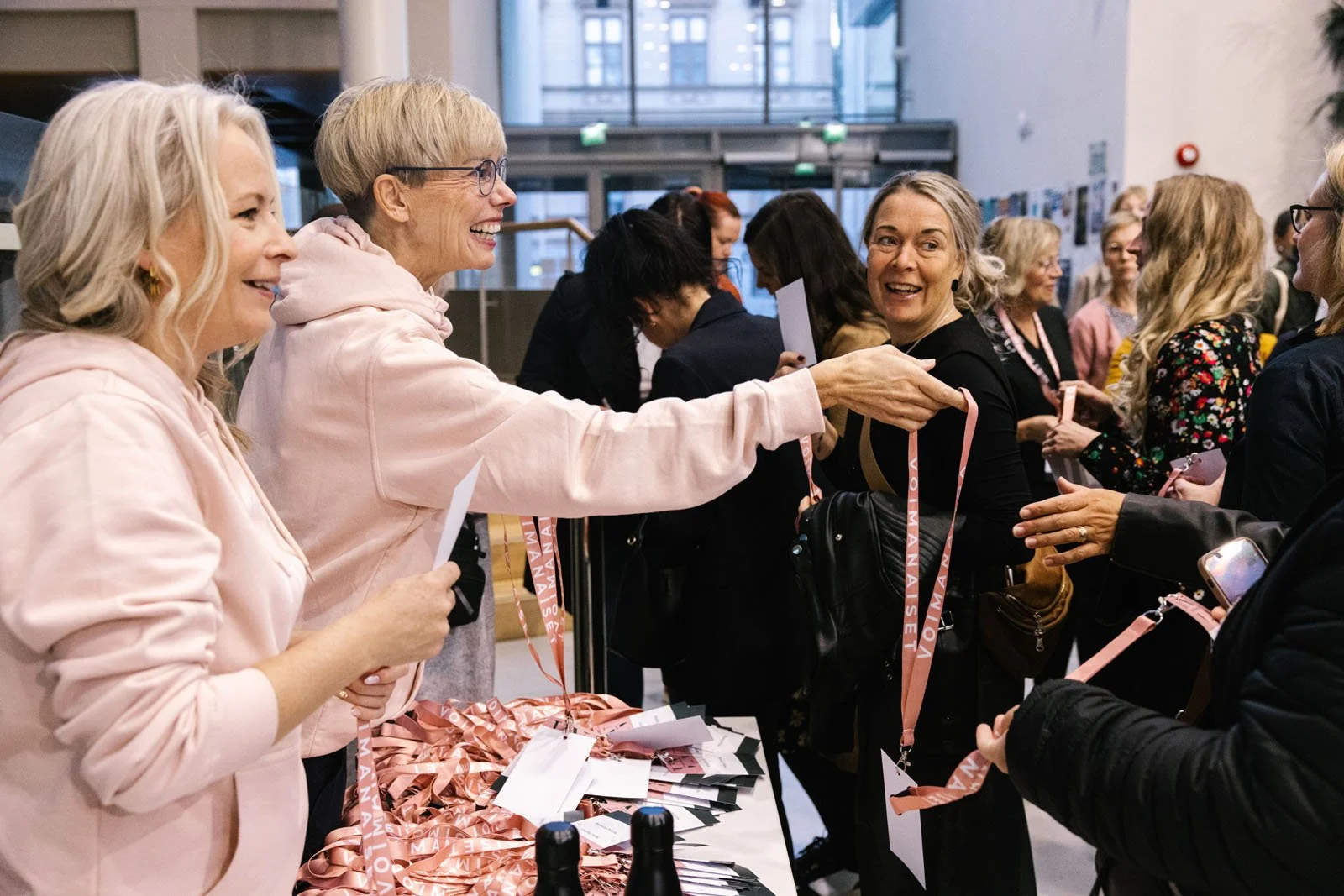 Naiset juttelevat ja hymyilevät tapahtuman rekisteritiskillä, jossa on roikkuvia nimilappuja ja lippiksiä.