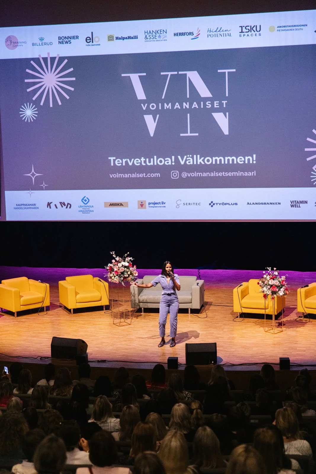 Naistele kannettava mikrofonia käyttäen esiintyjä lavalla, jossa on neljä keltaista ja yksi harmaa sohva sekä kaksi kukkalaatikkoa. Taustalla suuri näyttö, jossa tekstiä ja logoja tapahtuman nimeltä Voimanaiset, suomalaisista yrityksistä ja järjestöi