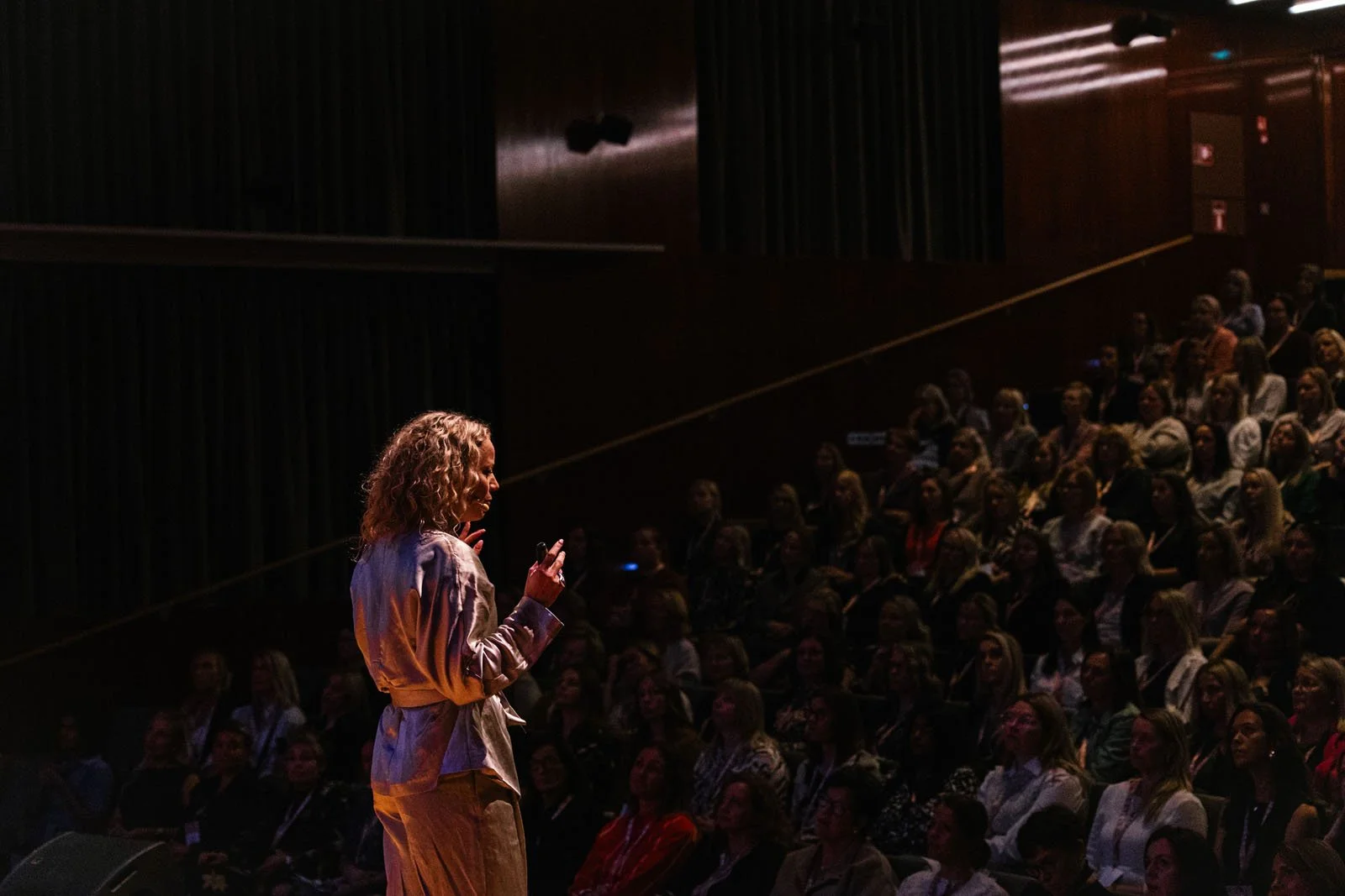 Nainen puhuu tai esittää, katse kohti näyttämöä, taustalla suuri ihmisjoukko istuu katsomossa, kaikki ovat vastavalossa.