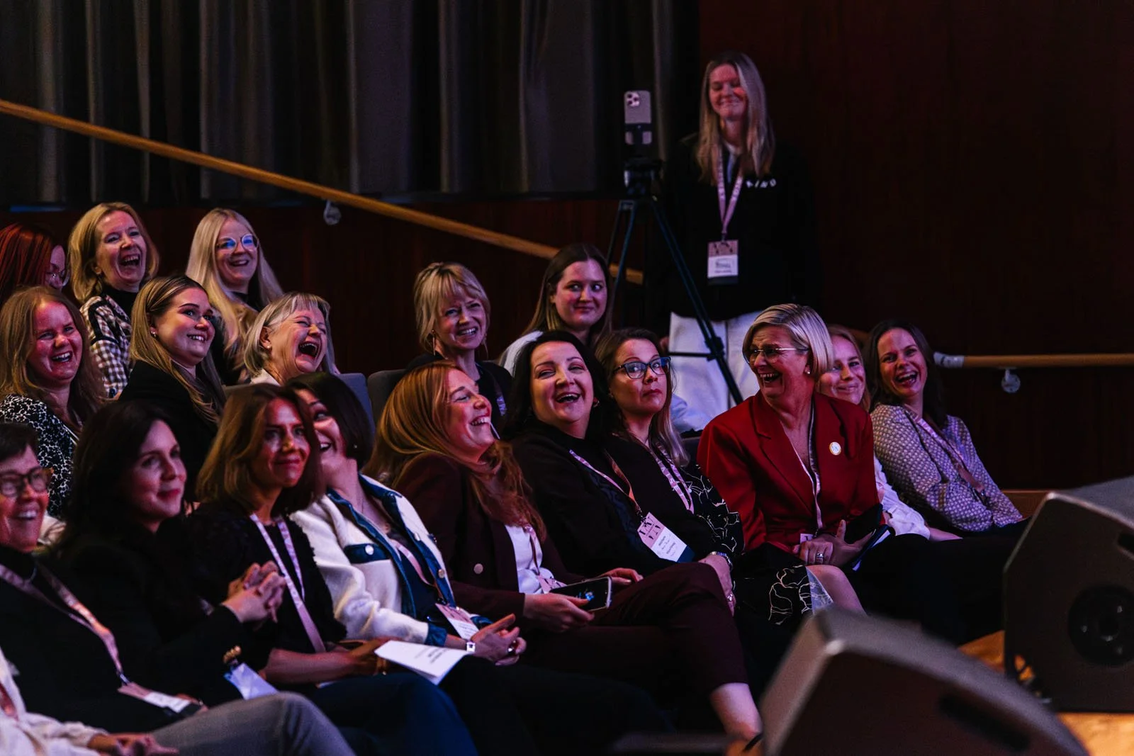 Lämminhenkinen ryhmä ihmisiä istuu ja nauraa konferenssissa tai tapahtumassa, sisätiloissa, pääasiassa eturivissä.