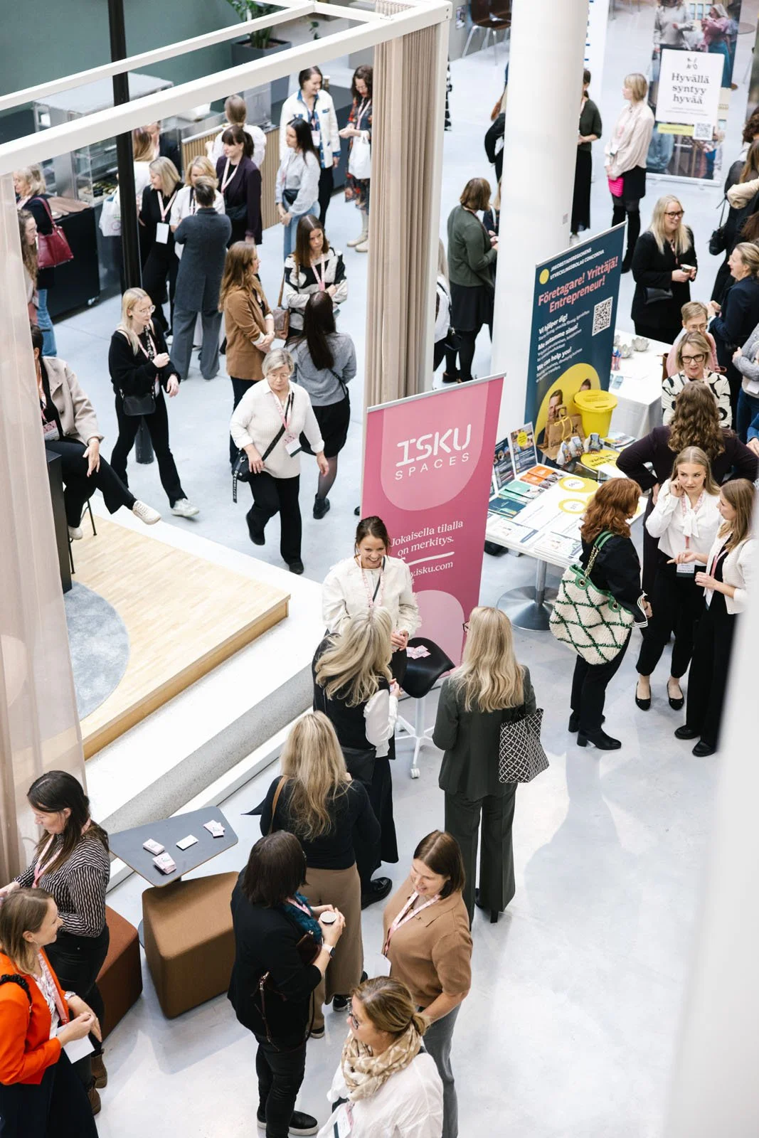 Tapahtumatilaisuus, jossa monia ihmisiä keskustelemassa ja osallistumassa erilaisiin toimintoihin. Näkyvissä on esittelypisteitä ja rekkikojuja, joissa mainostetaan ISKU SEINÄÄ ja muuta yritysmarkkinointia.