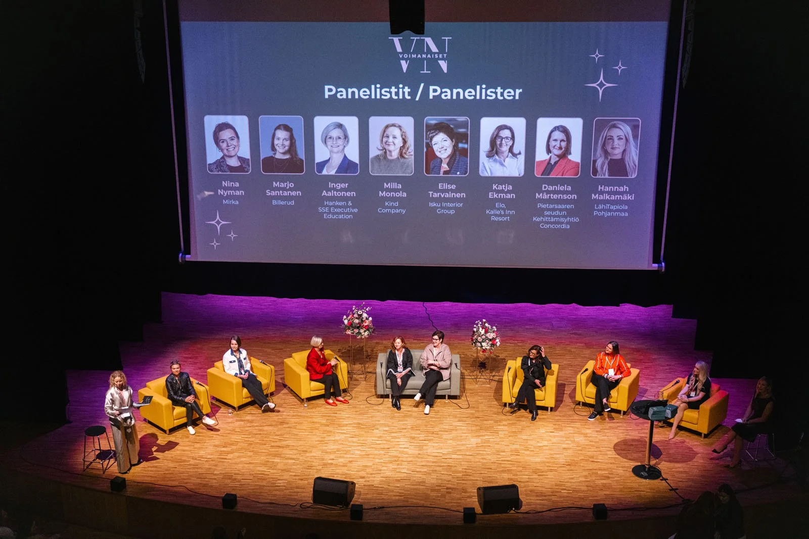 Keskellä lavalla on useita ihmisiä istumassa nojatuoleissa, vieressä kukkalaareja. Taustalla suuri näyttöruutu esittää puheillisten nimet ja kuvat. Yleisö katsoo tapahtumaa.