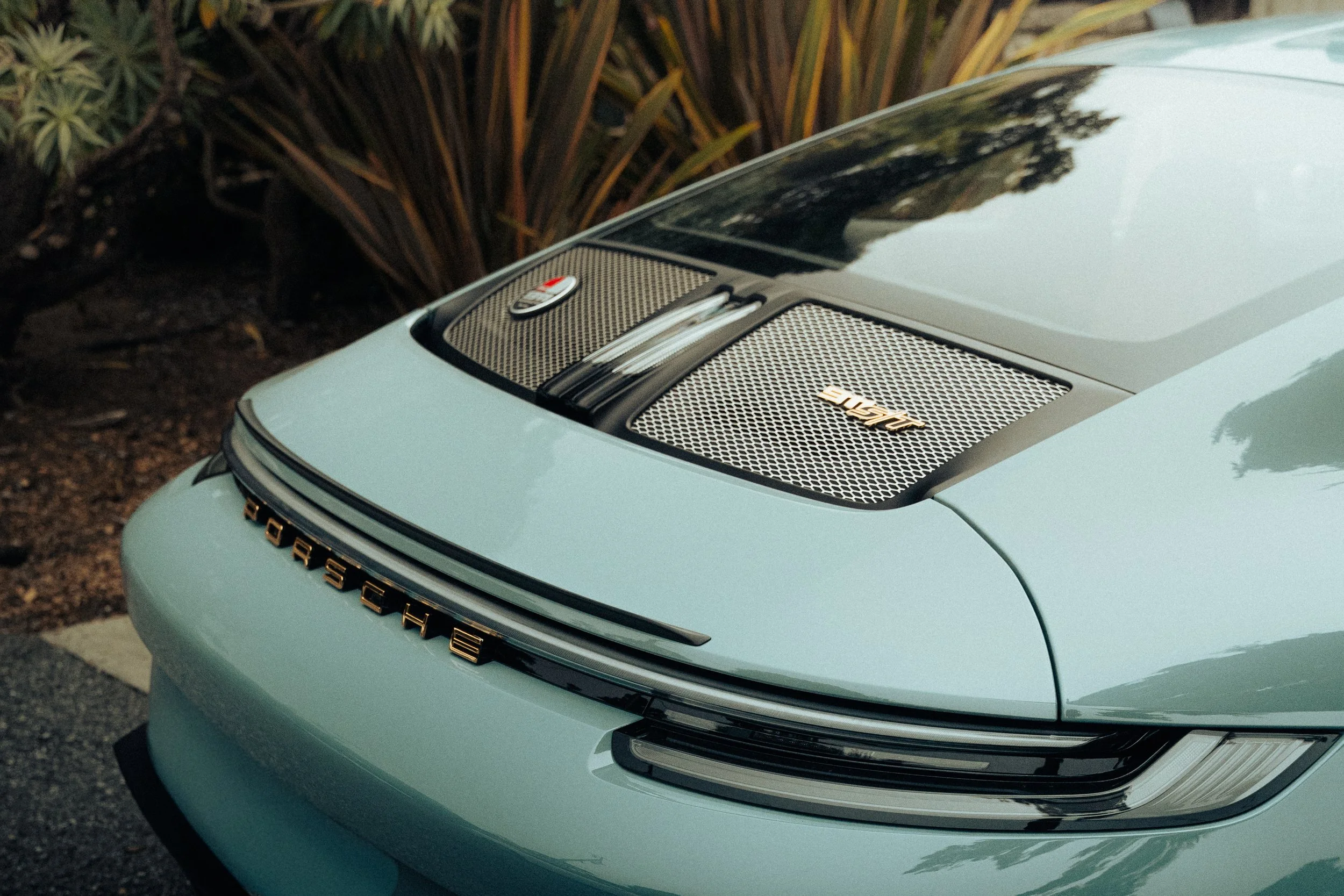 Close-up of the front of a light blue Porsche car showing the Porsche logo on the hood and a high-performance air intake grille.