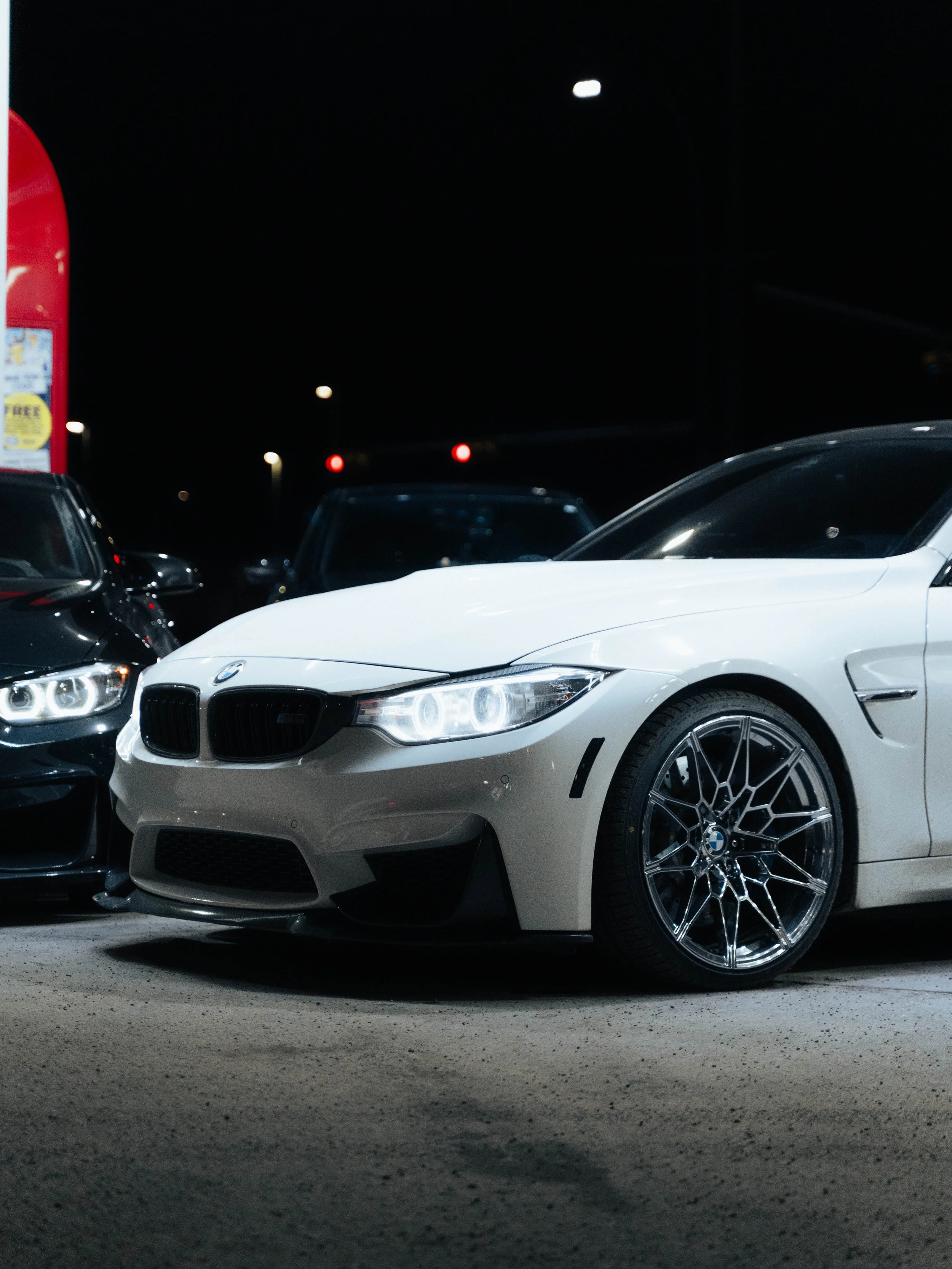 A white BMW sports car with black and chrome wheels parked in a lot at night, with neighboring cars and a red parking lot sign visible.