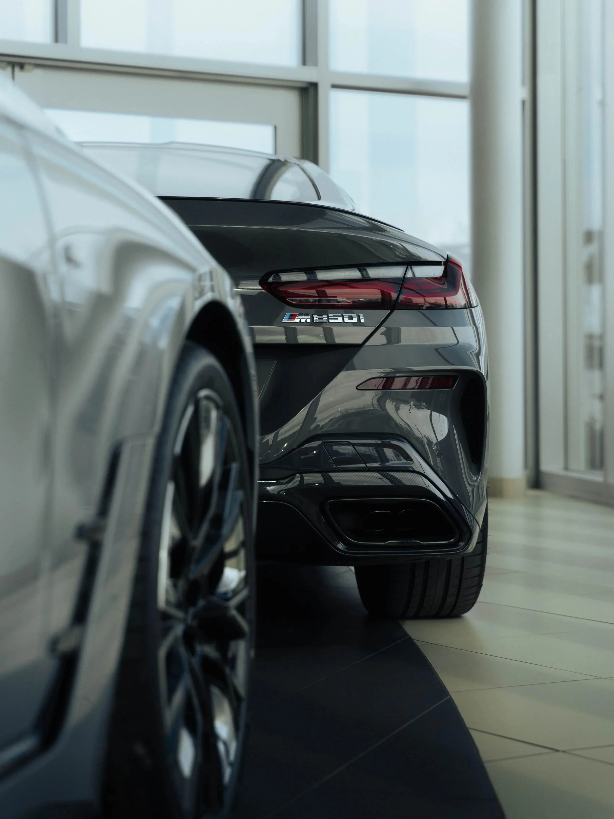 The image shows a close-up view of the rear ends of two cars parked indoors, with one being a dark gray BMW M850i, model badge visible, and the other a silver vehicle visible only partially.