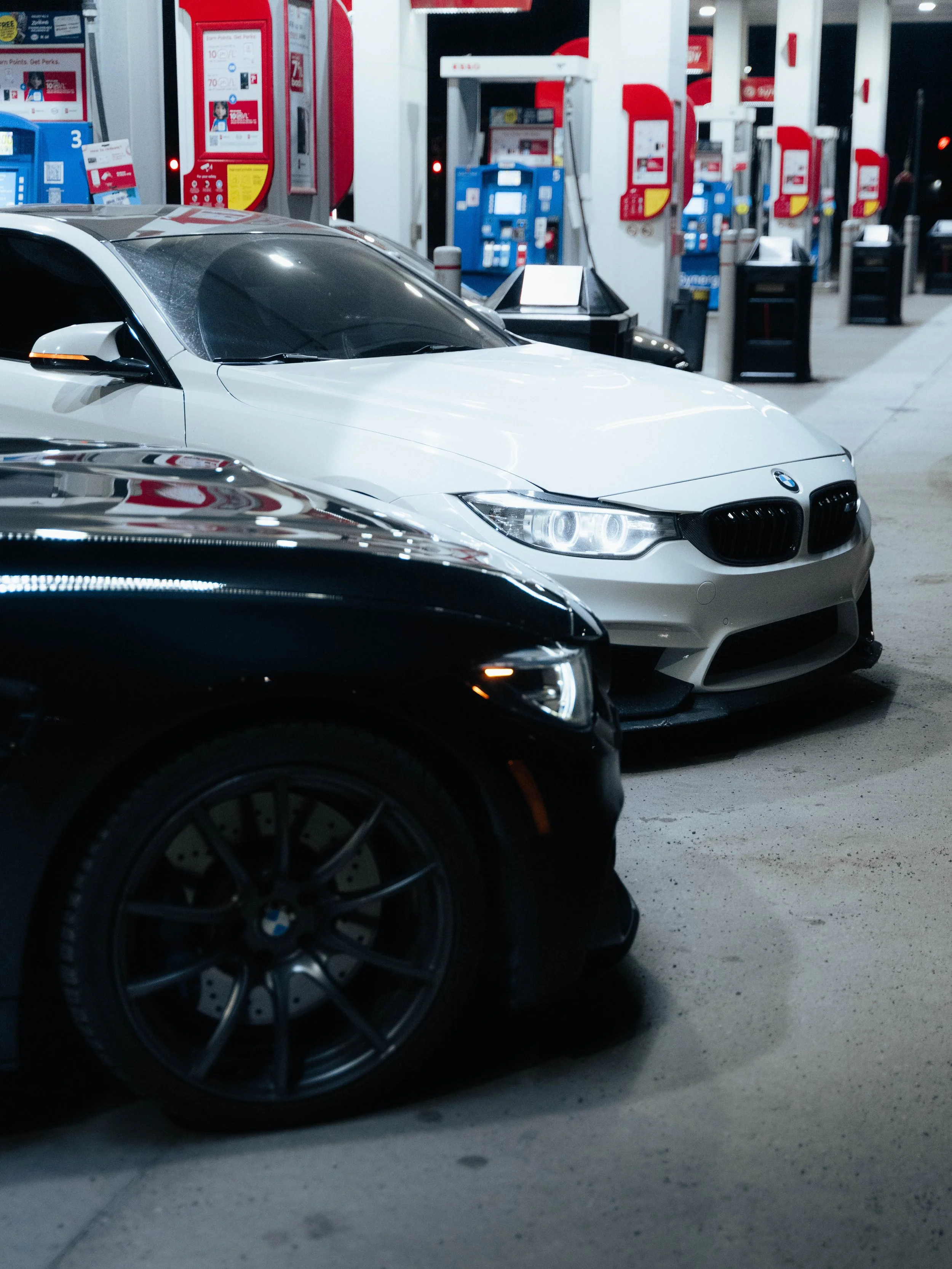 A row of parked cars at a gas station during nighttime. The cars include a black vehicle in the foreground and a white BMW behind it. The gas station has red and blue fuel pumps, with some showing digital screens and card readers.