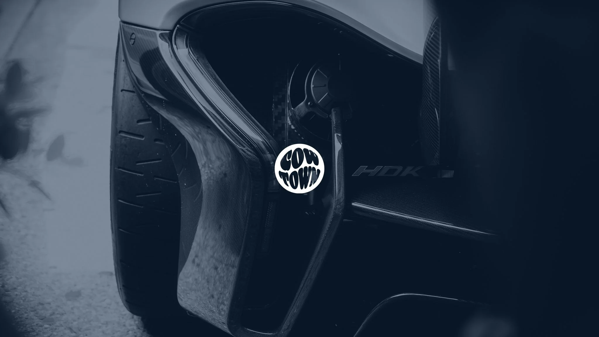 Close-up of the front corner of a black luxury car, showing the headlight and tire, with Cowtown Media logo.