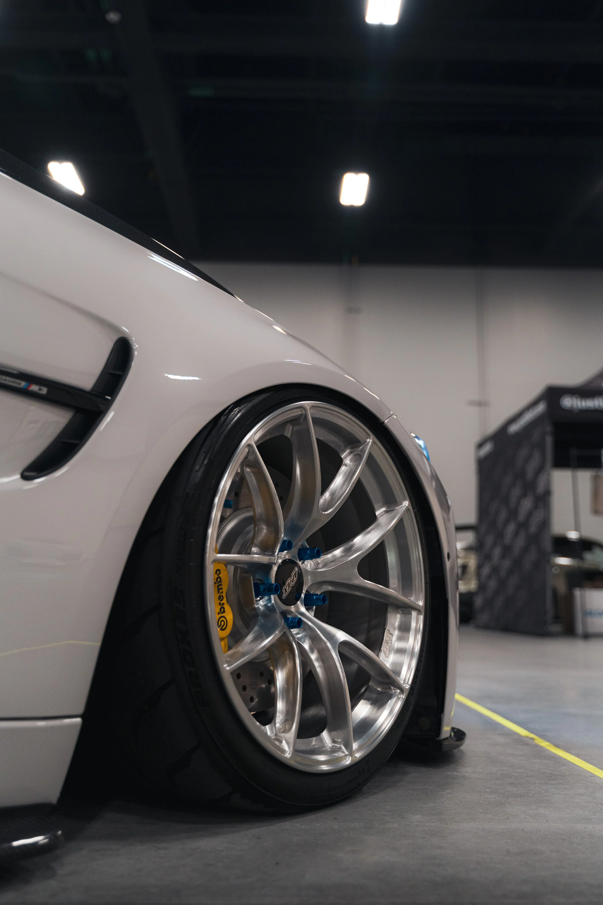 Close-up of a white race car's front left tire and wheel, with a yellow brake caliper visible, in an indoor garage or exhibition space.