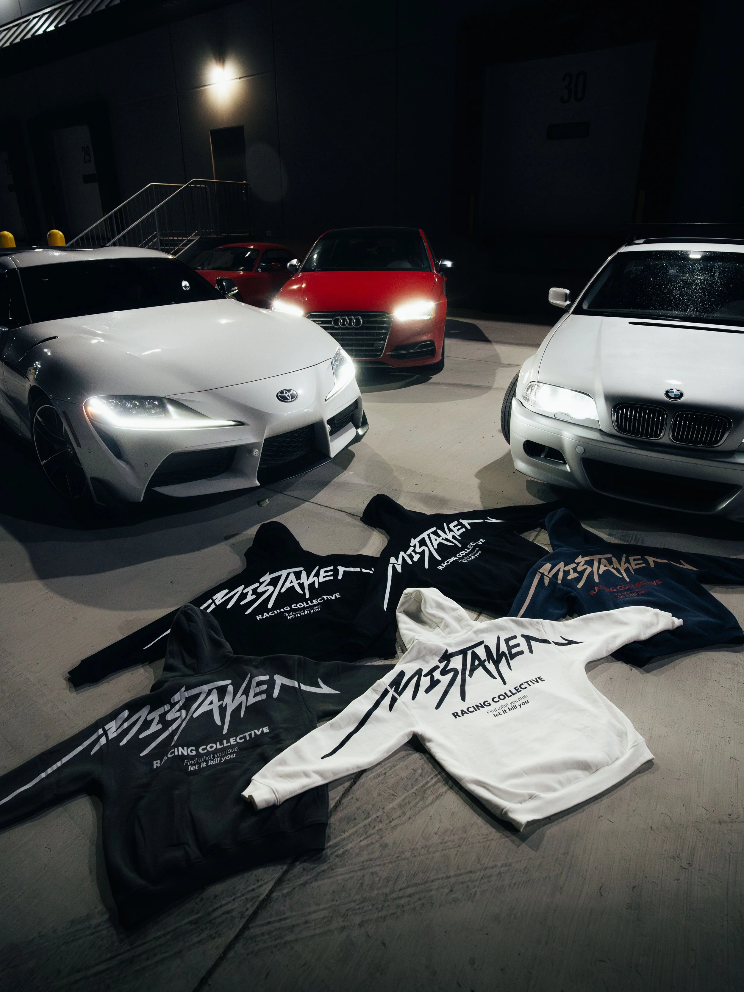 Several hoodies with 'MISTAKEN RACING COLLECTIVE' logo laid out on the ground in front of parked cars, including a white Toyota, a red Audi, and a white BMW, in a dimly lit parking garage.
