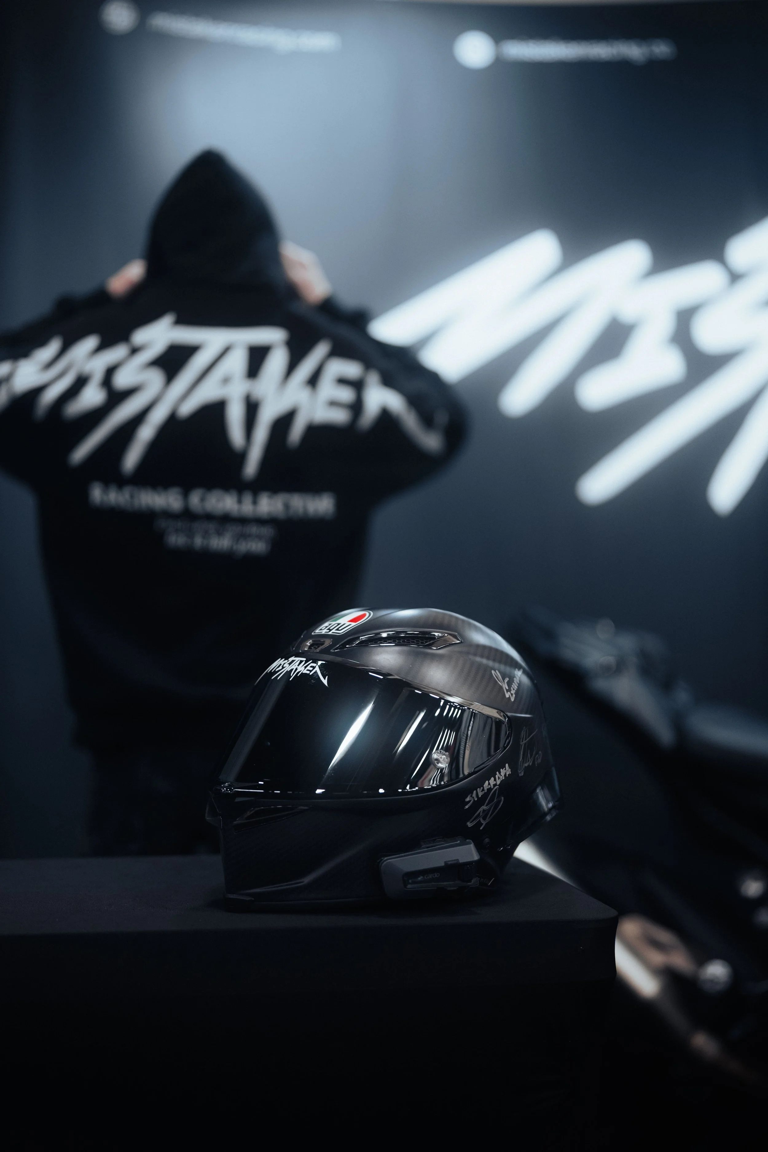 A black motorcycle helmet with reflective visor and white branding, placed on a black surface, with a person in a black hoodie in the background in front of a dark wall with Mistaken Racing Collective.
