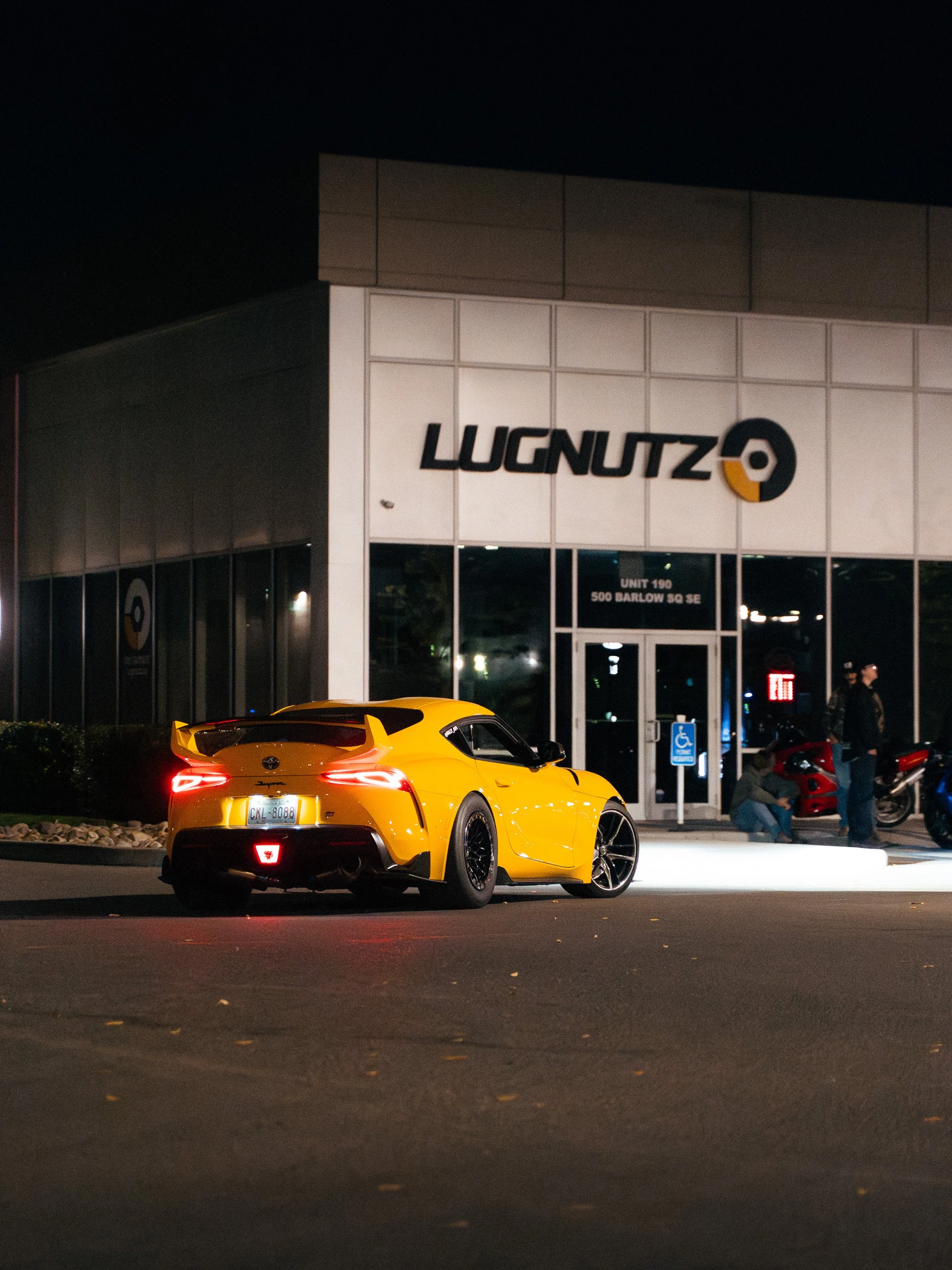 A yellow sports car parked outside a building at night, with people near the entrance.