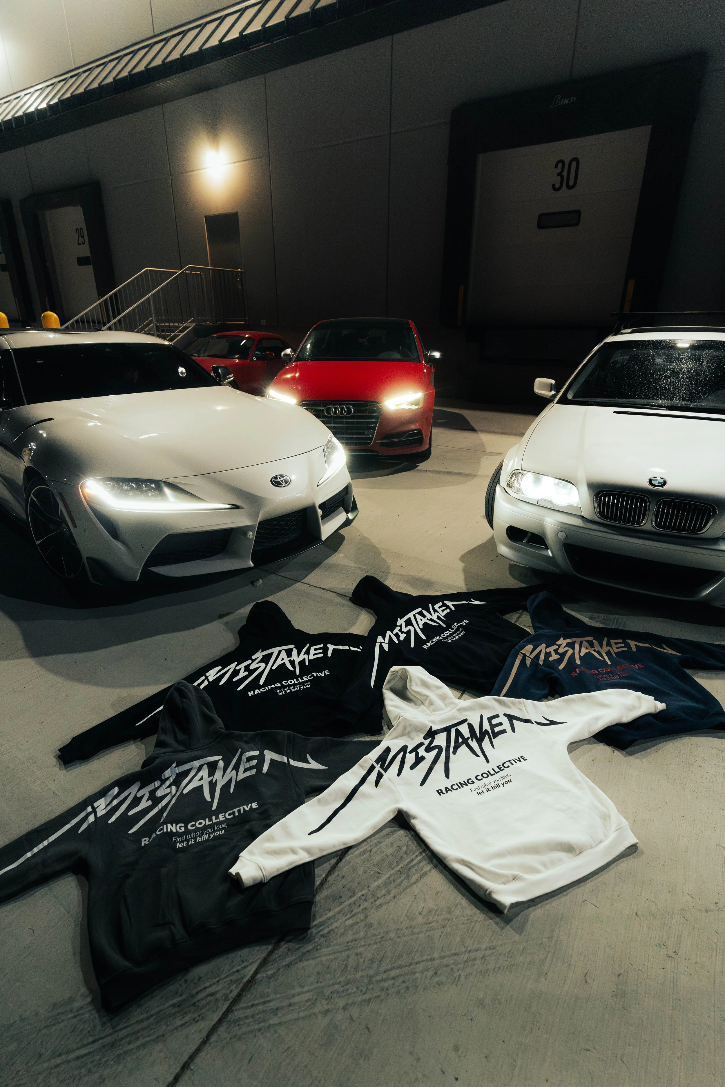 Multiple cars parked in a garage with hoodies laid out on the floor in front, displaying the text 'Mistaken Racing Collective.'
