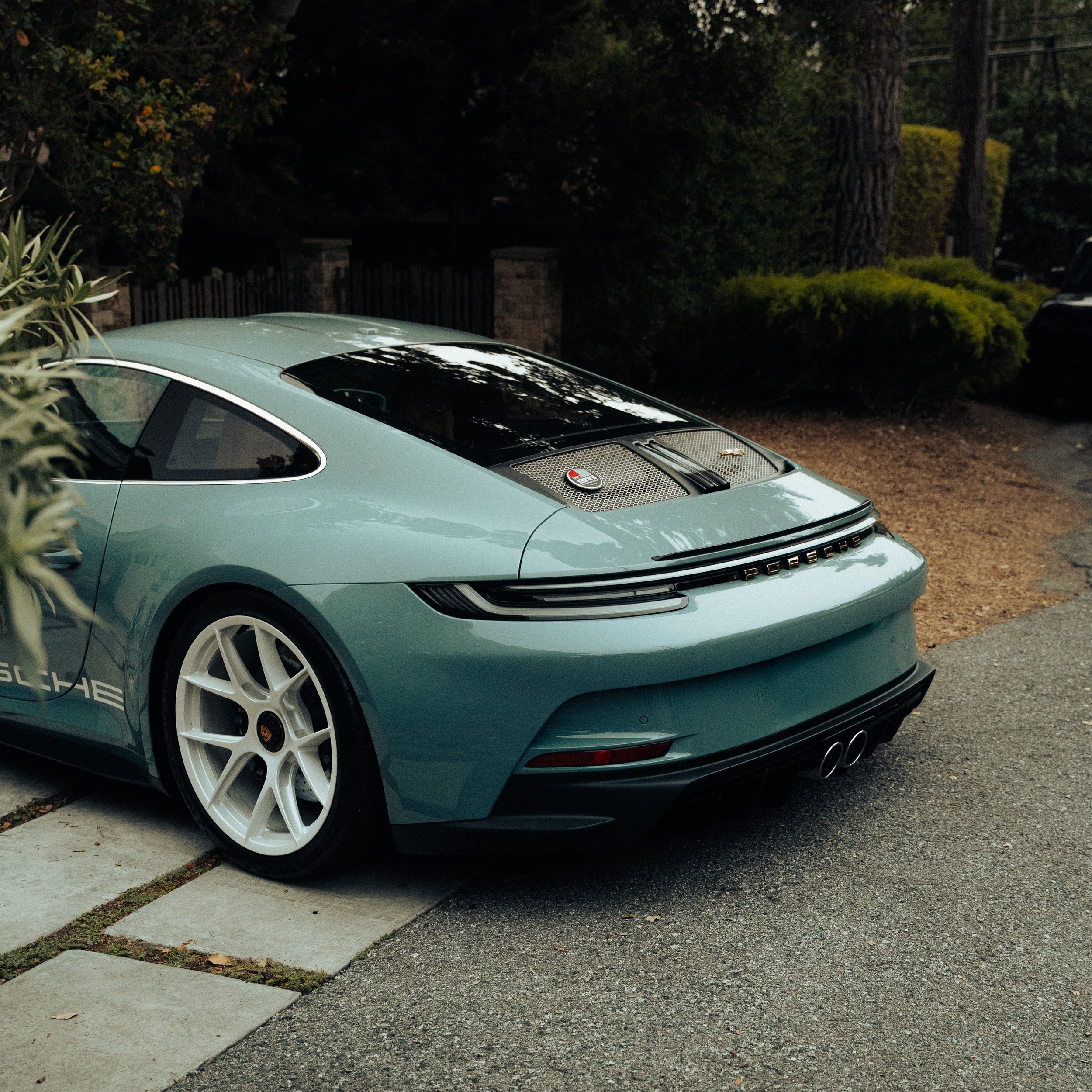 A light blue Porsche sports car parked on a driveway with greenery and trees in the background.