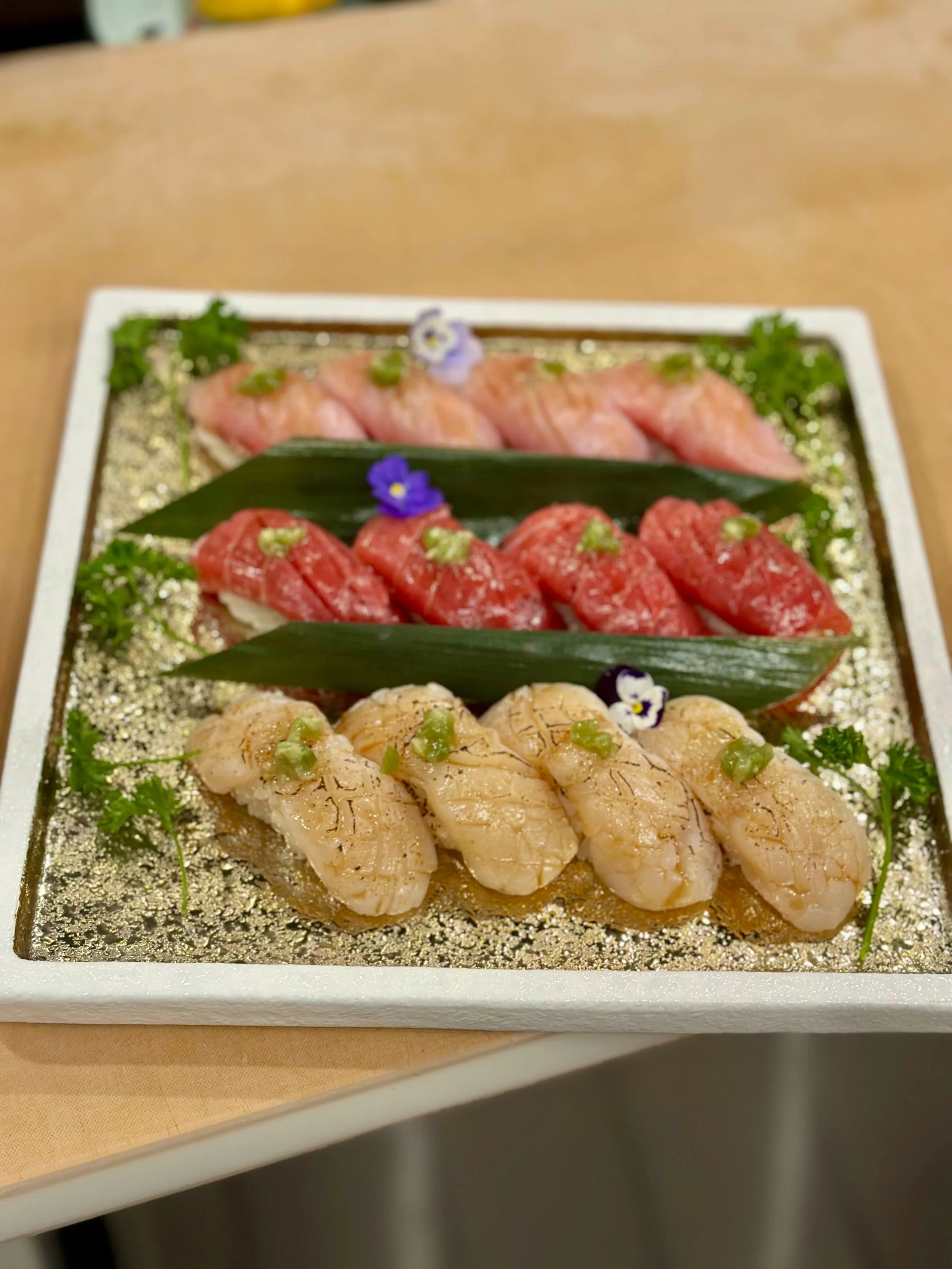 A decorative sushi platter featuring assorted nigiri sushi arranged on a white plate with gold accents, topped with garnish and flowers.