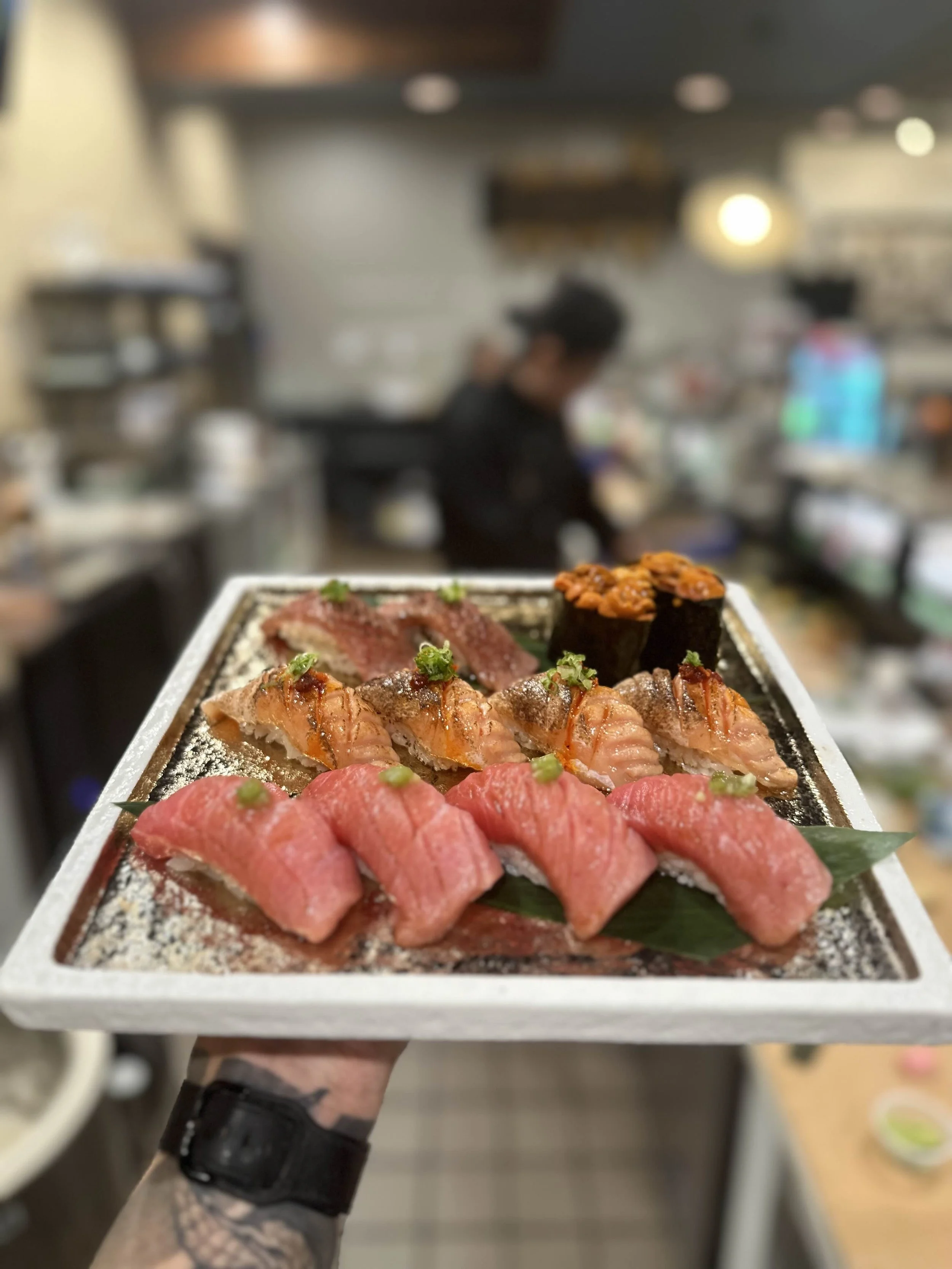 Assorted sushi platter with various types of sushi, including tuna, salmon, and sea urchin, presented on a plate held by a tattooed arm, with a blurred restaurant background.