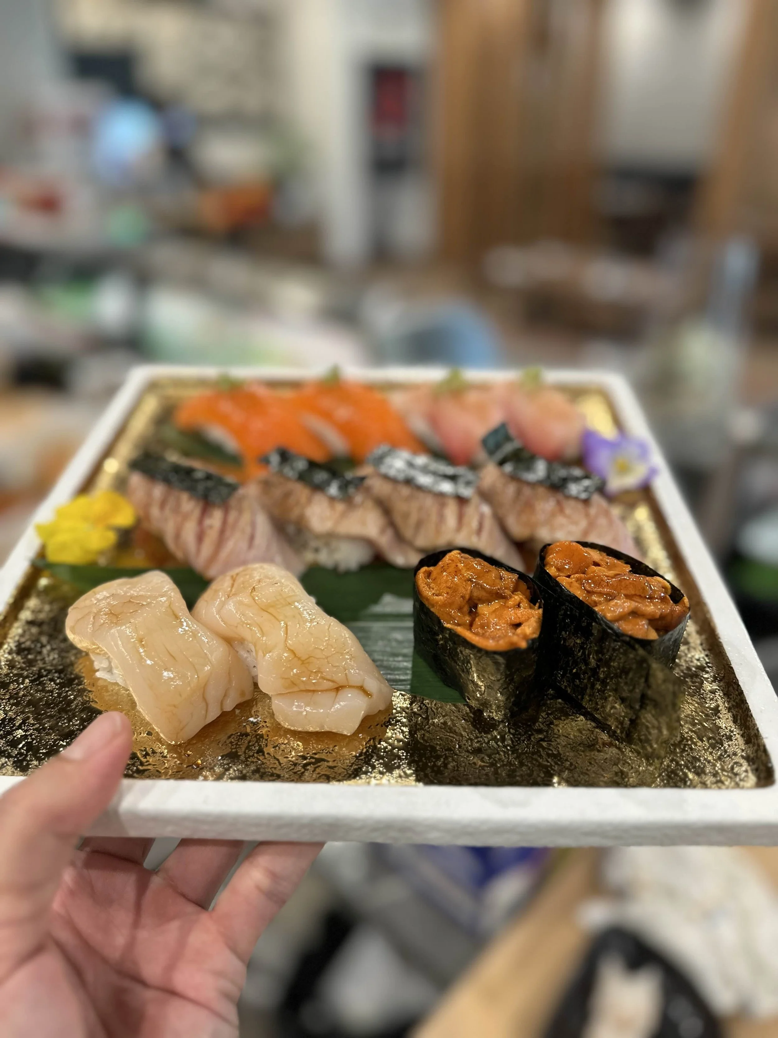 A close-up of a sushi platter with a variety of sushi pieces including nigiri and gunkan. The platter includes scallop nigiri, salmon nigiri, and sea urchin gunkan, garnished with flowers. The background is blurred.