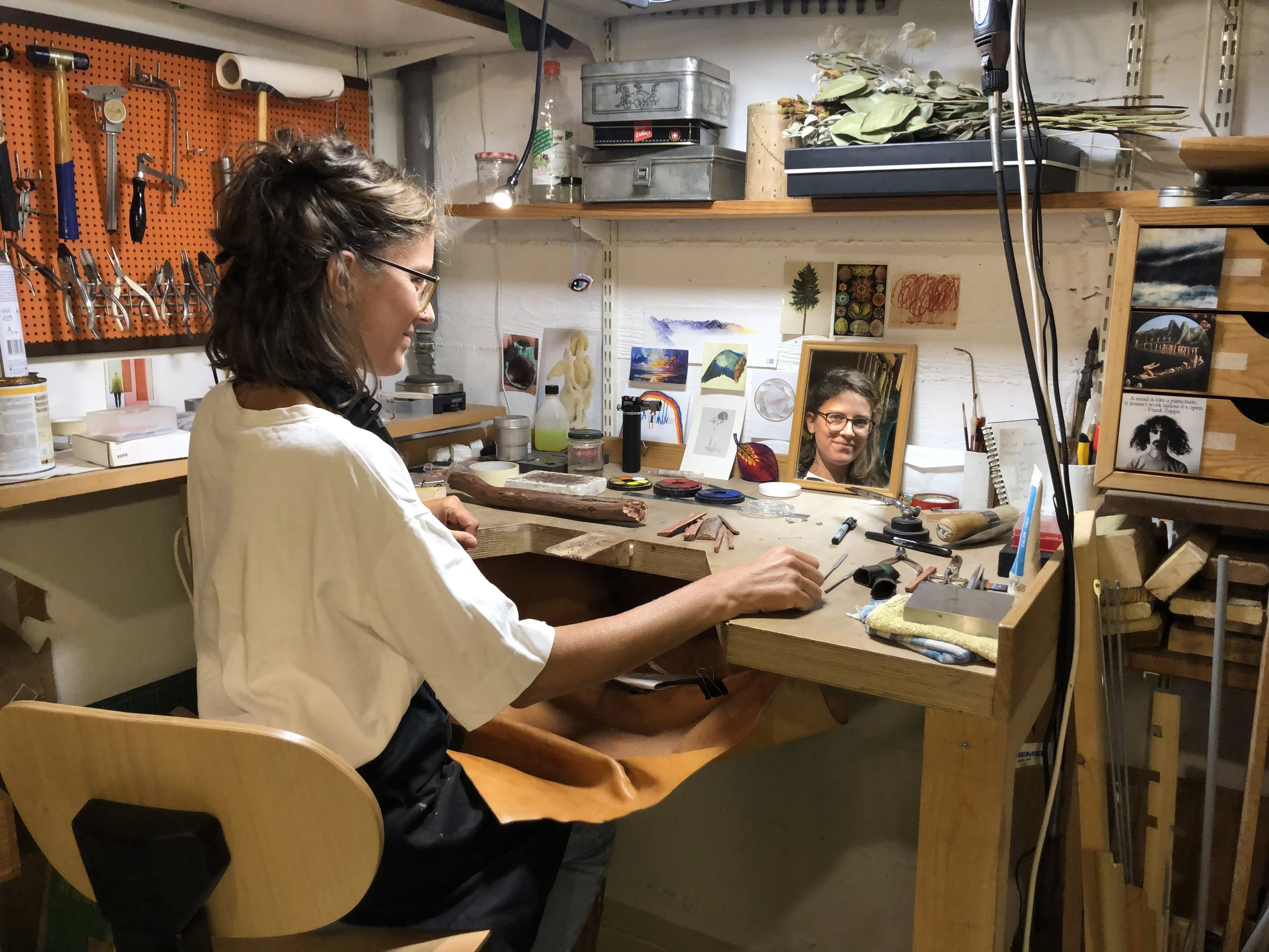 A woman working at a jewelry-making bench with tools, a mirror reflecting her face, and art supplies, in a well-organized workshop.