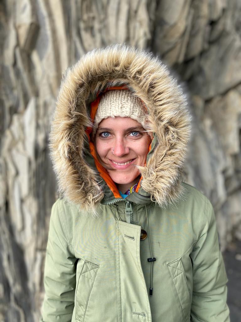 Young woman in a green winter coat with a hood lined with faux fur, wearing a beige knit beanie, smiling, standing in front of a stone wall.