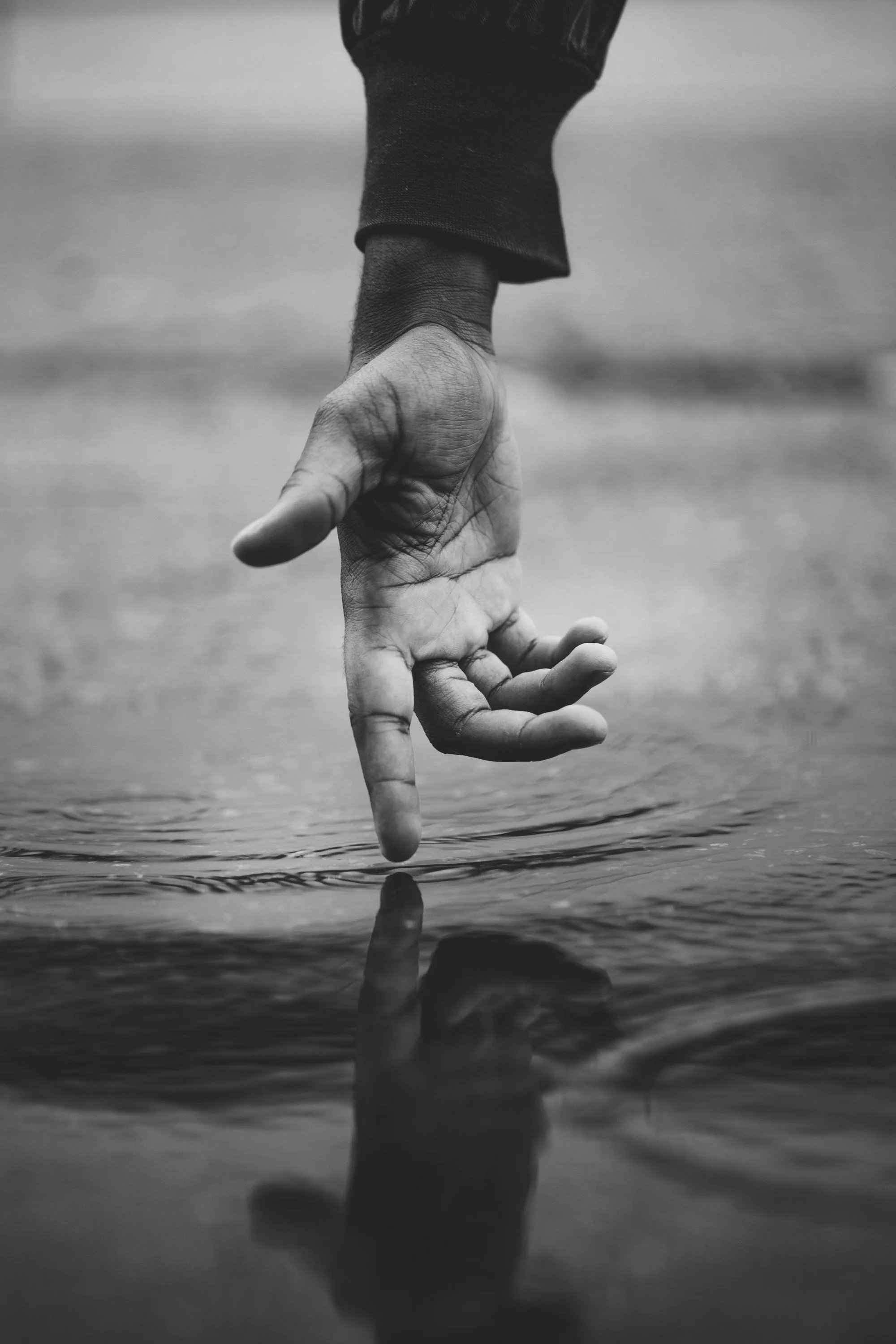 A black and white photo of a hand with the index finger touching water, creating ripples.