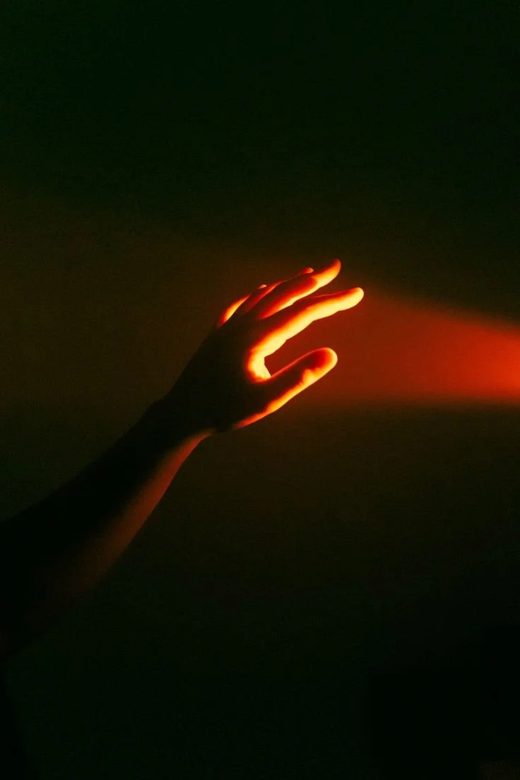 A hand reaching into darkness with a red light shining through the fingers.