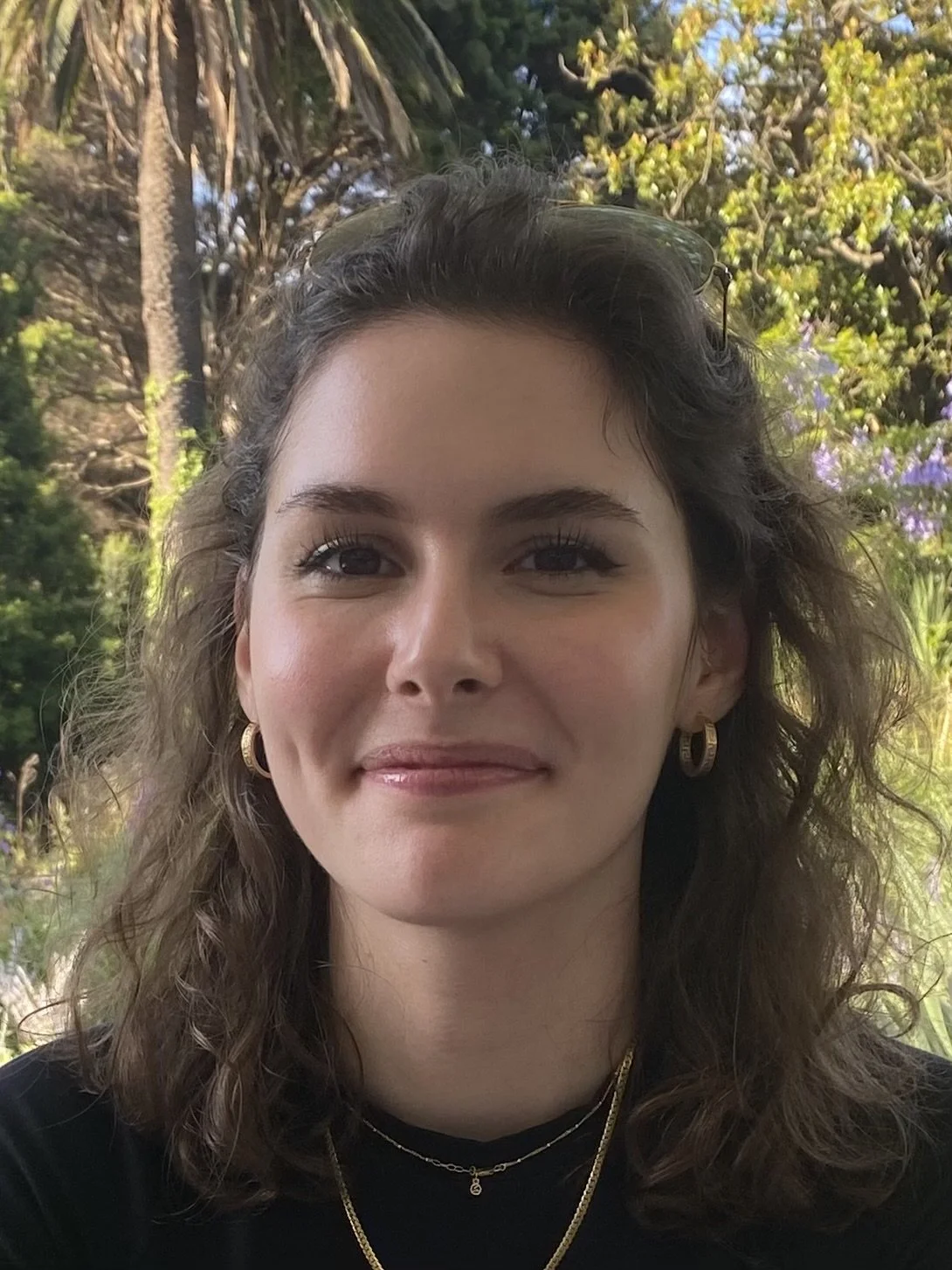 Close-up photo of a woman with wavy brown hair, wearing gold hoop earrings, layered gold necklaces, and a black top, outdoors in a lush garden with trees and purple flowers in the background.