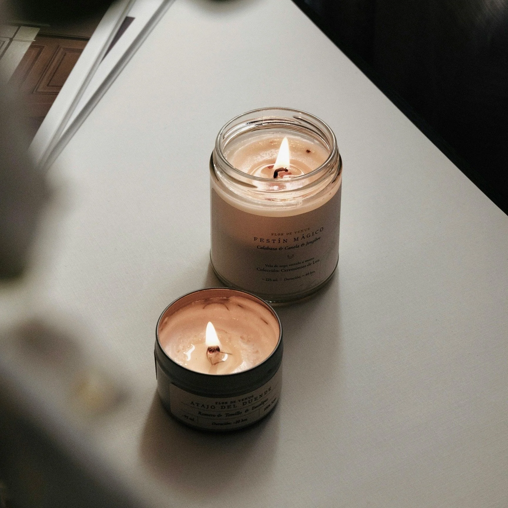 Two lit scented candles in glass jars on a table, casting a warm glow.
