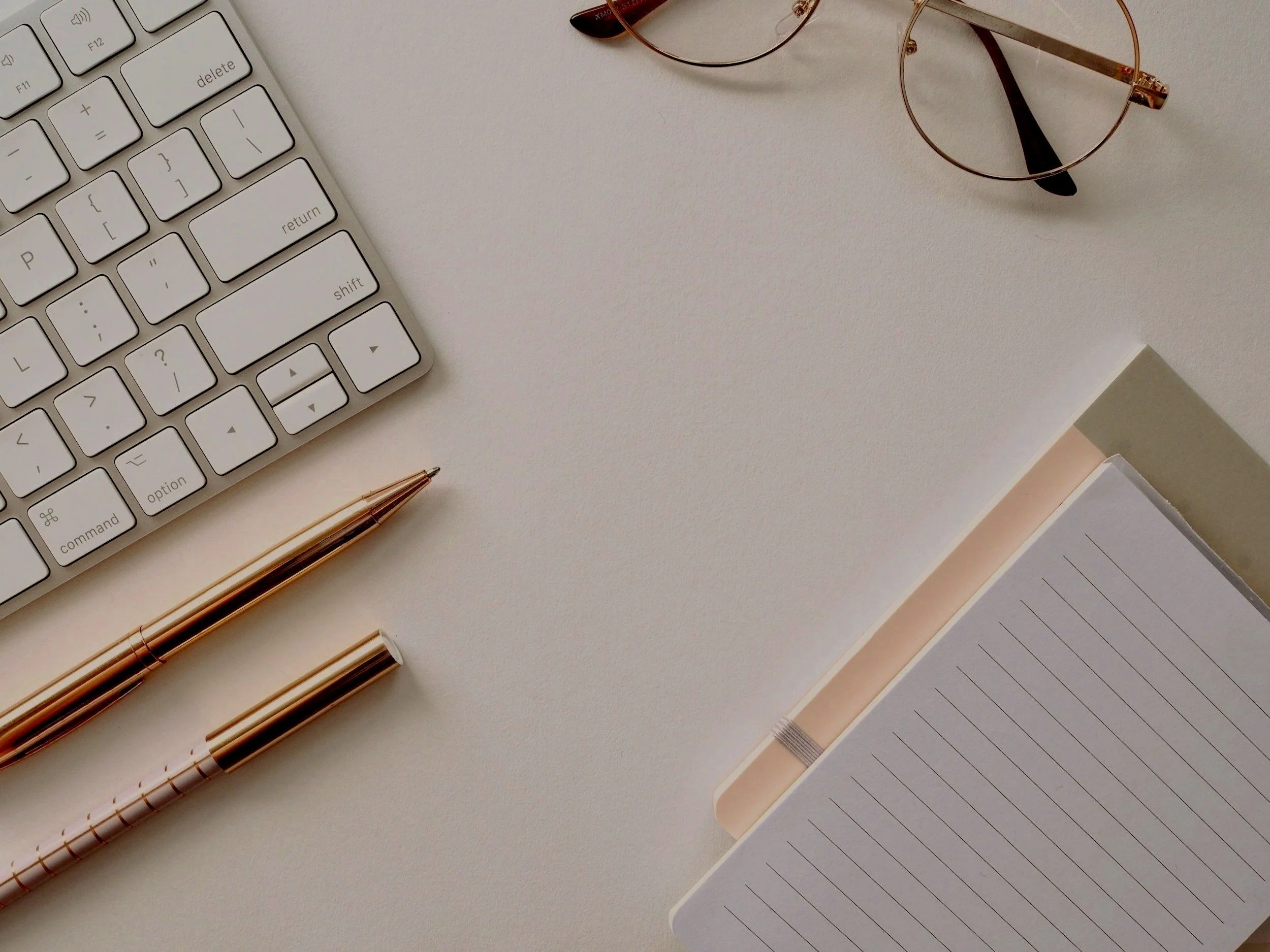 Overhead view of a keyboard, two pens, glasses, and a lined notebook on a beige surface.