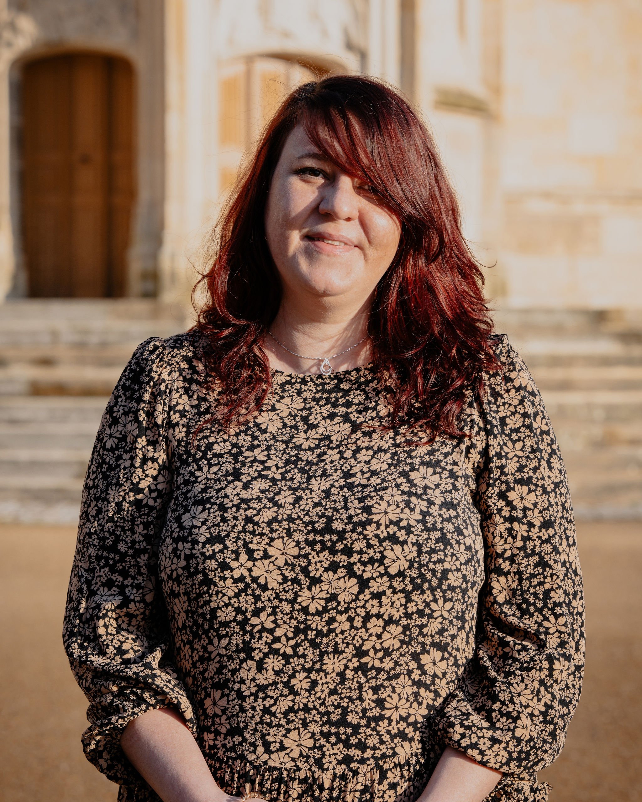 Femme avec cheveux roux foncé en robe à motifs de fleurs, devant un bâtiment en pierre en plein jour.