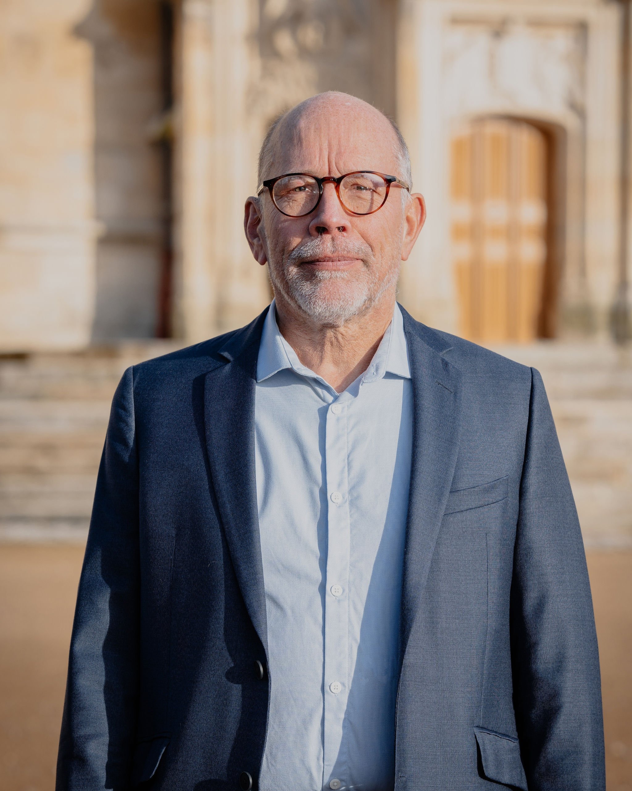 Homme âgé portant des lunettes, en costume bleu, devant un bâtiment ancien en pierre.
