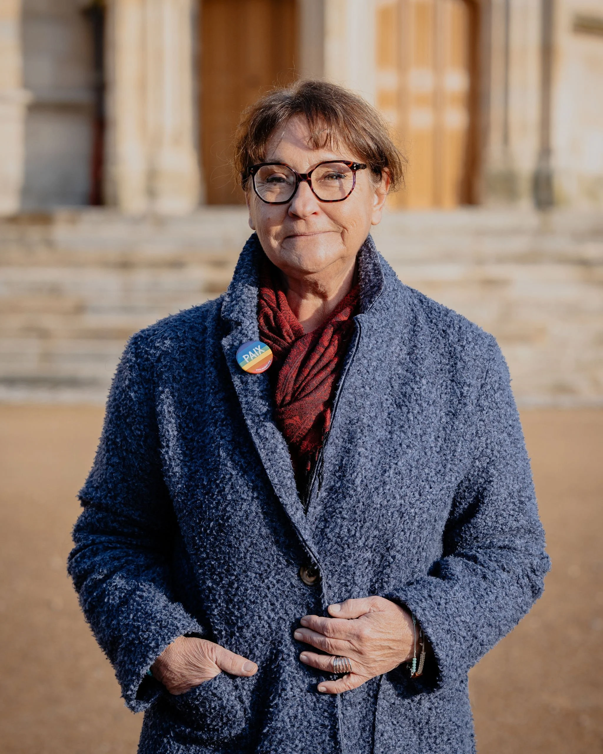Une femme âgée avec des lunettes, portant un manteau bleu et un foulard rouge, pose devant une façade en pierre. Elle porte un badge avec le mot 'PAIX'.