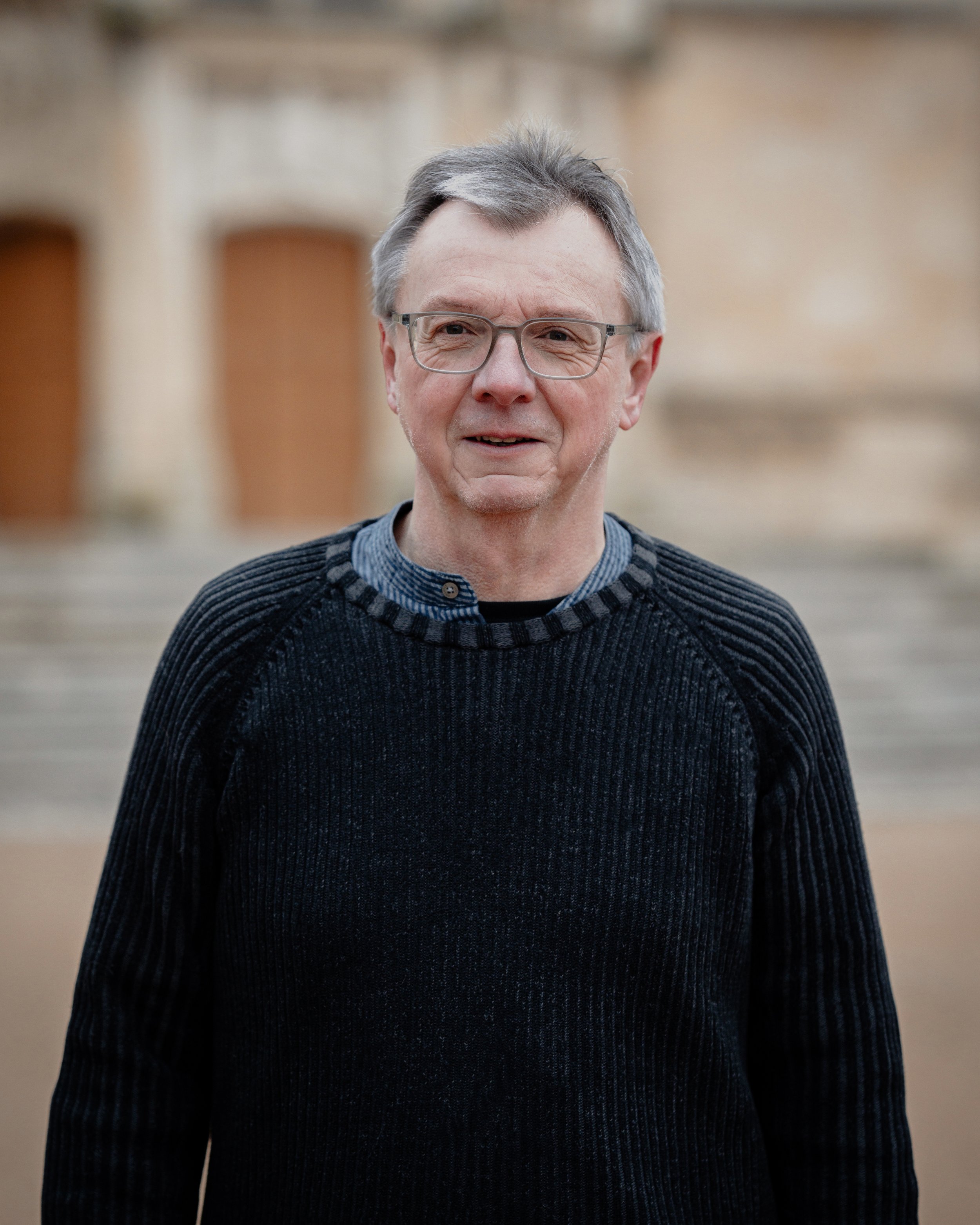 Un homme âgé portant des lunettes et un pull noir, debout à l'extérieur, avec des bâtiments flous en arrière-plan.