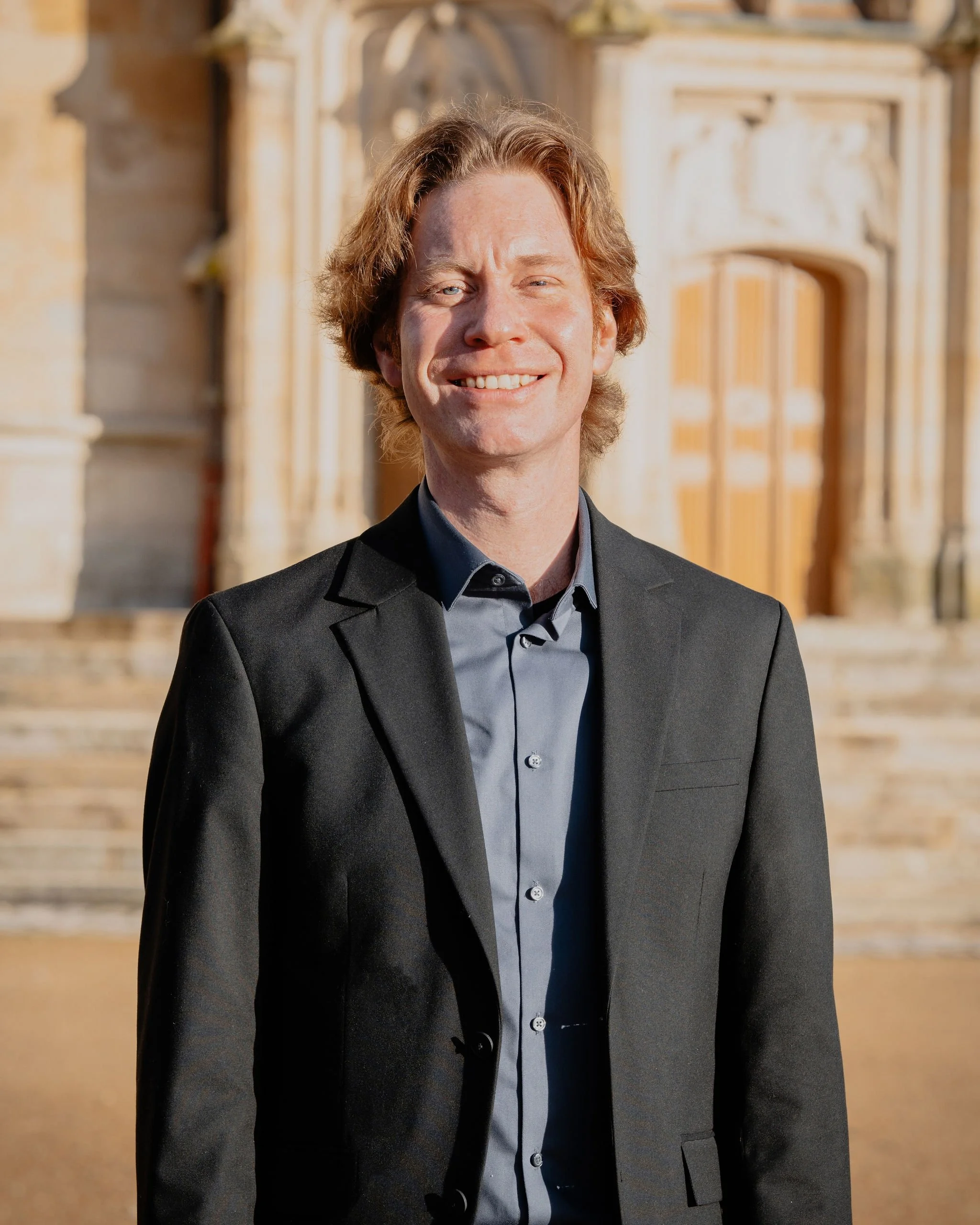 Un homme souriant en costume noir, debout devant un bâtiment ancien en pierre, architecture historique, en plein jour.