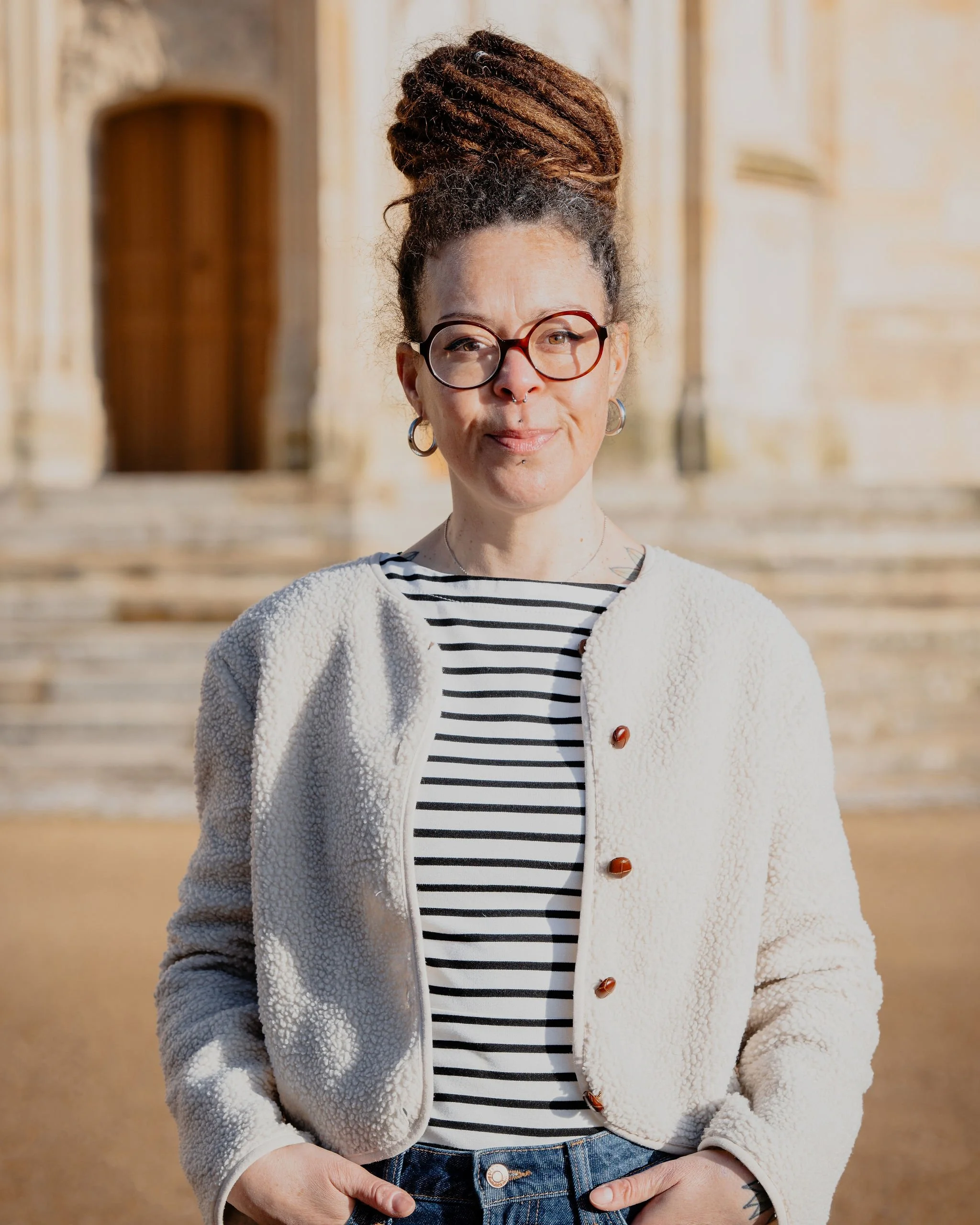 Jeune femme à dreadlocks portant des lunettes, un jean et une veste en tissu doucement bouclé, debout devant un bâtiment ancien en pierre.