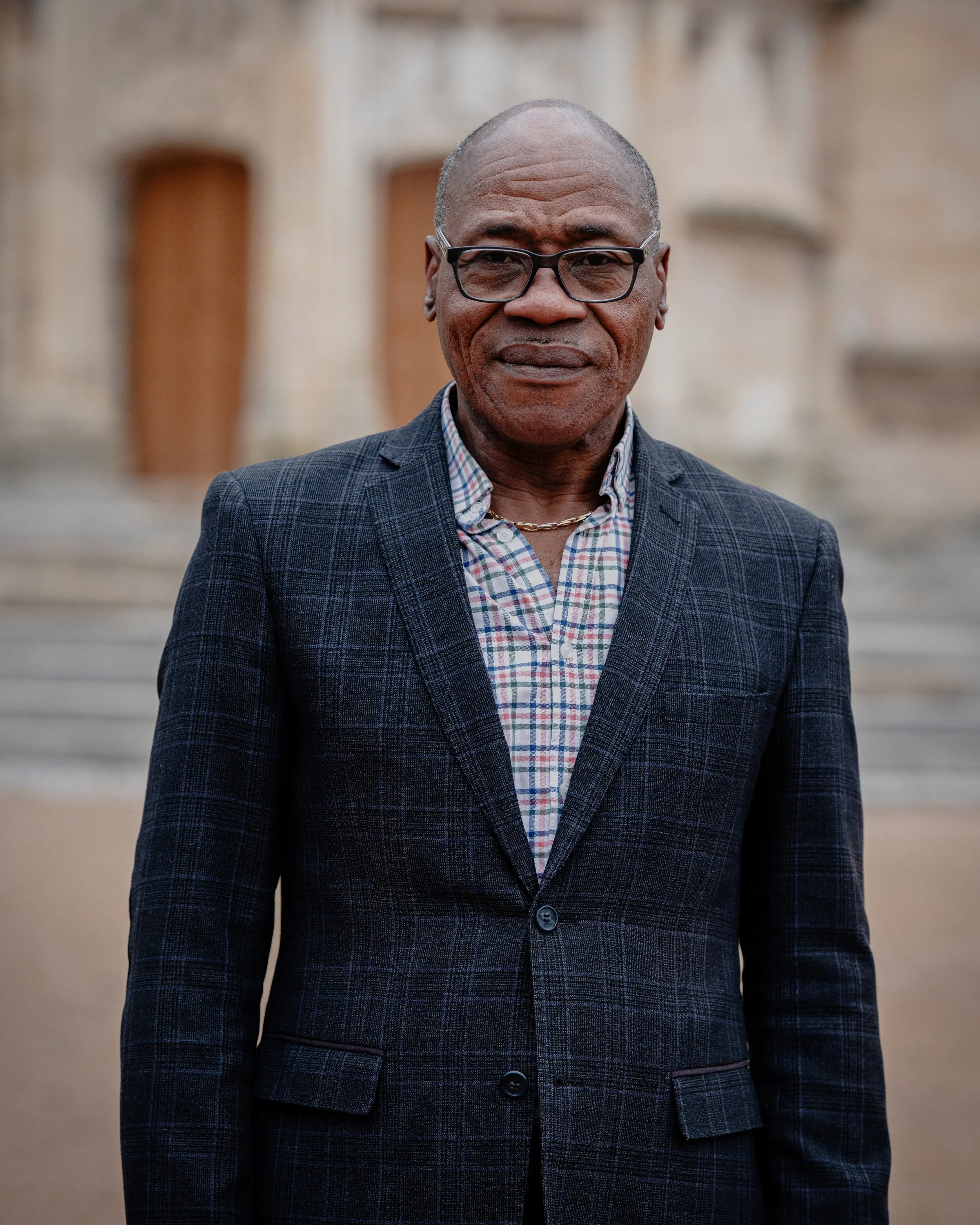 Un homme âgé portant des lunettes, une veste à carreaux et une chemise à motifs se tient devant un bâtiment ancien en pierre.