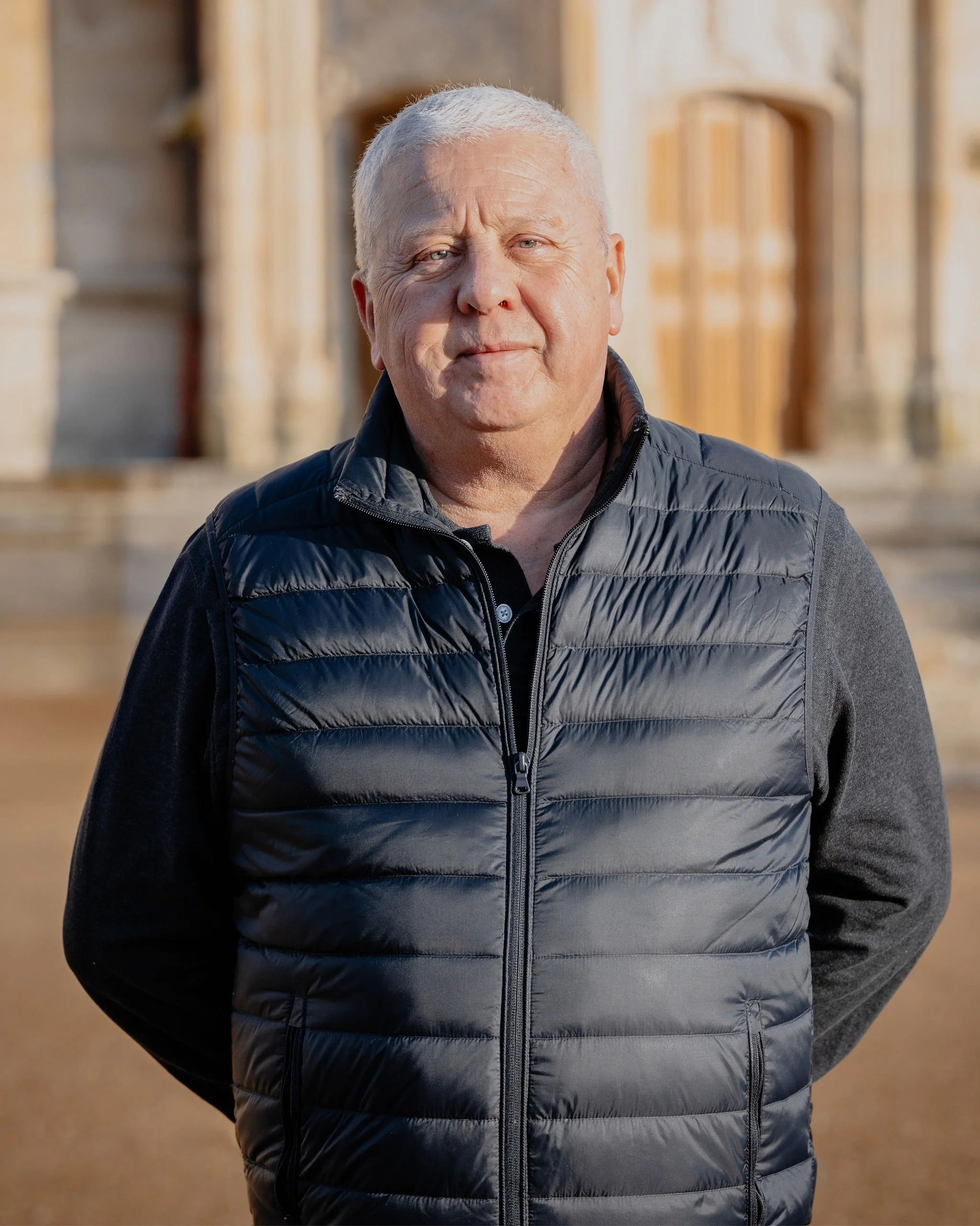 Un homme âgé avec des cheveux gris, portant un gilet noir matelassé et un pull gris foncé, pose devant un bâtiment en pierre, avec un léger sourire, lors d'une journée ensoleillée.