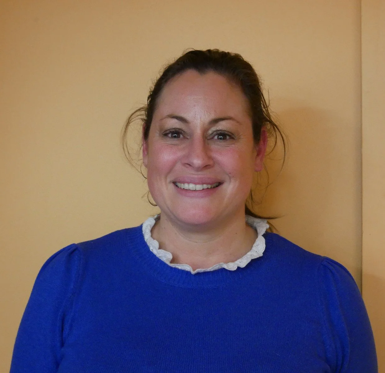 Une femme souriante portant un pull bleu avec un col blanc en dentelle, debout devant un mur beige.