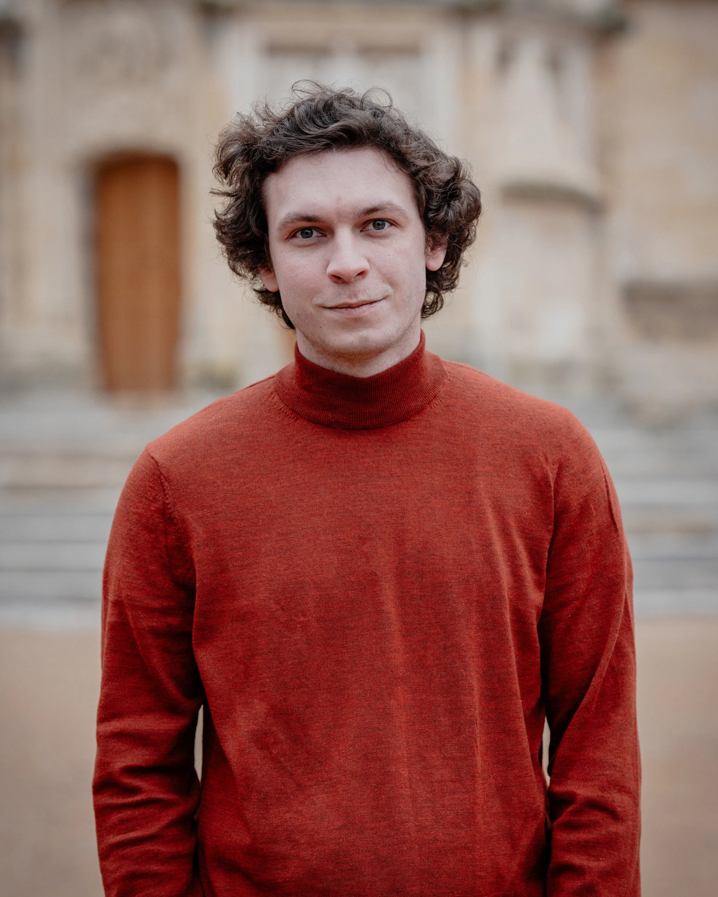 Jeune homme avec cheveux bouclés portant un pull rouge devant un bâtiment en pierre.