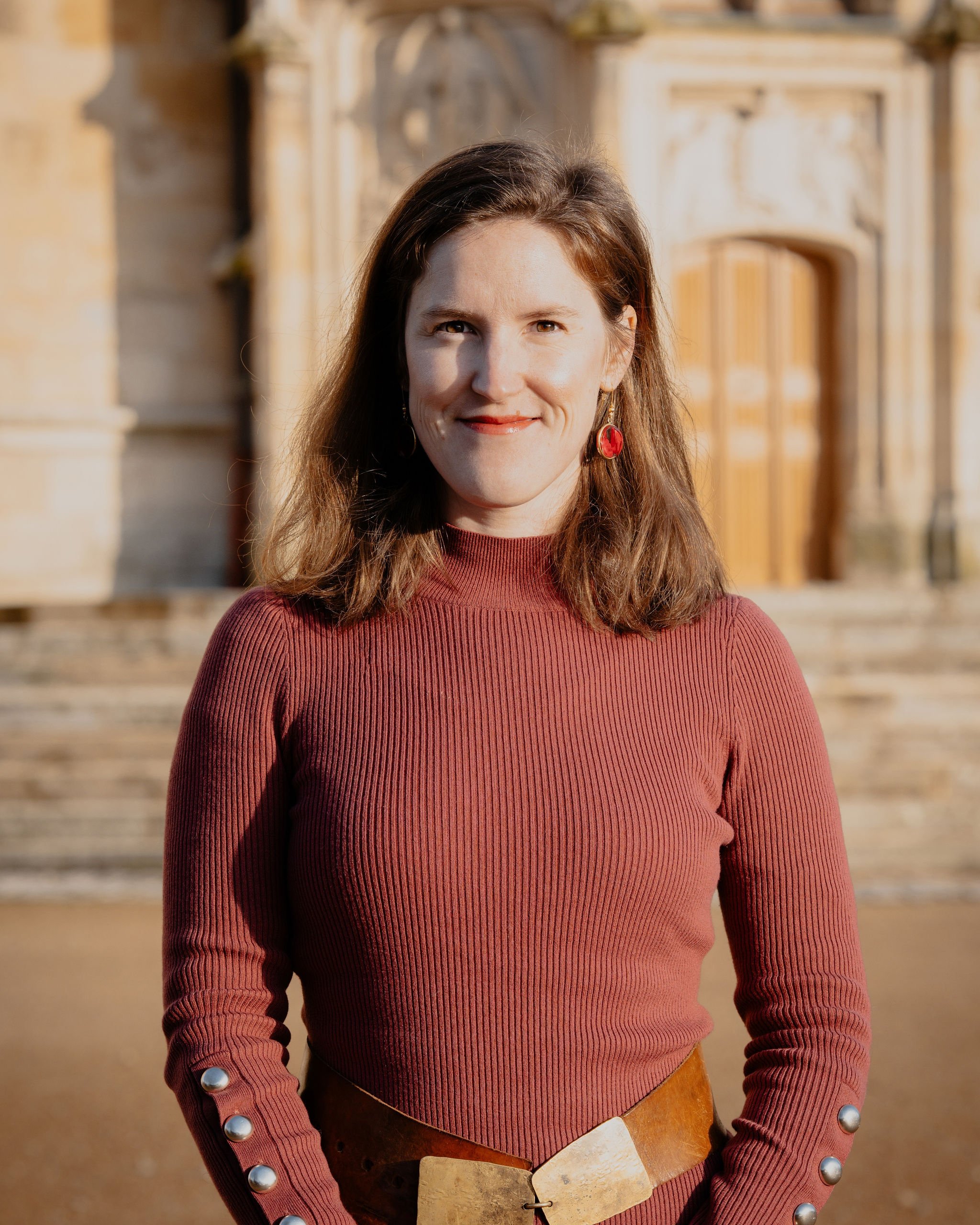 Une femme souriante avec des cheveux bruns, portant un pull rouge avec des boutons sur les manches, et des boucles d'oreilles rouges, devant un bâtiment en pierre ancien.