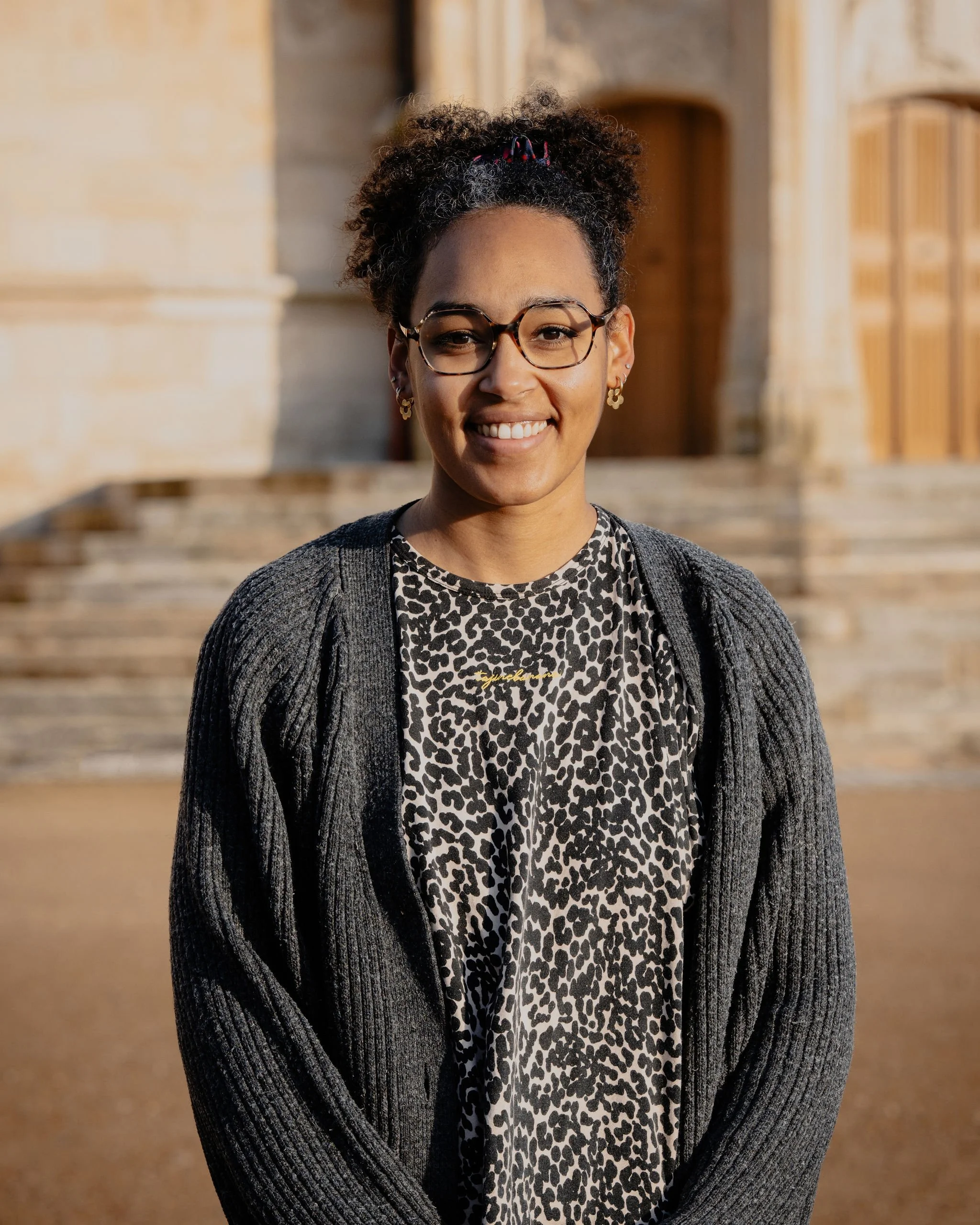 Une femme souriante portant des lunettes, un chemisier à motifs léopard, un gilet gris foncé, avec un bâtiment en pierre en arrière-plan.