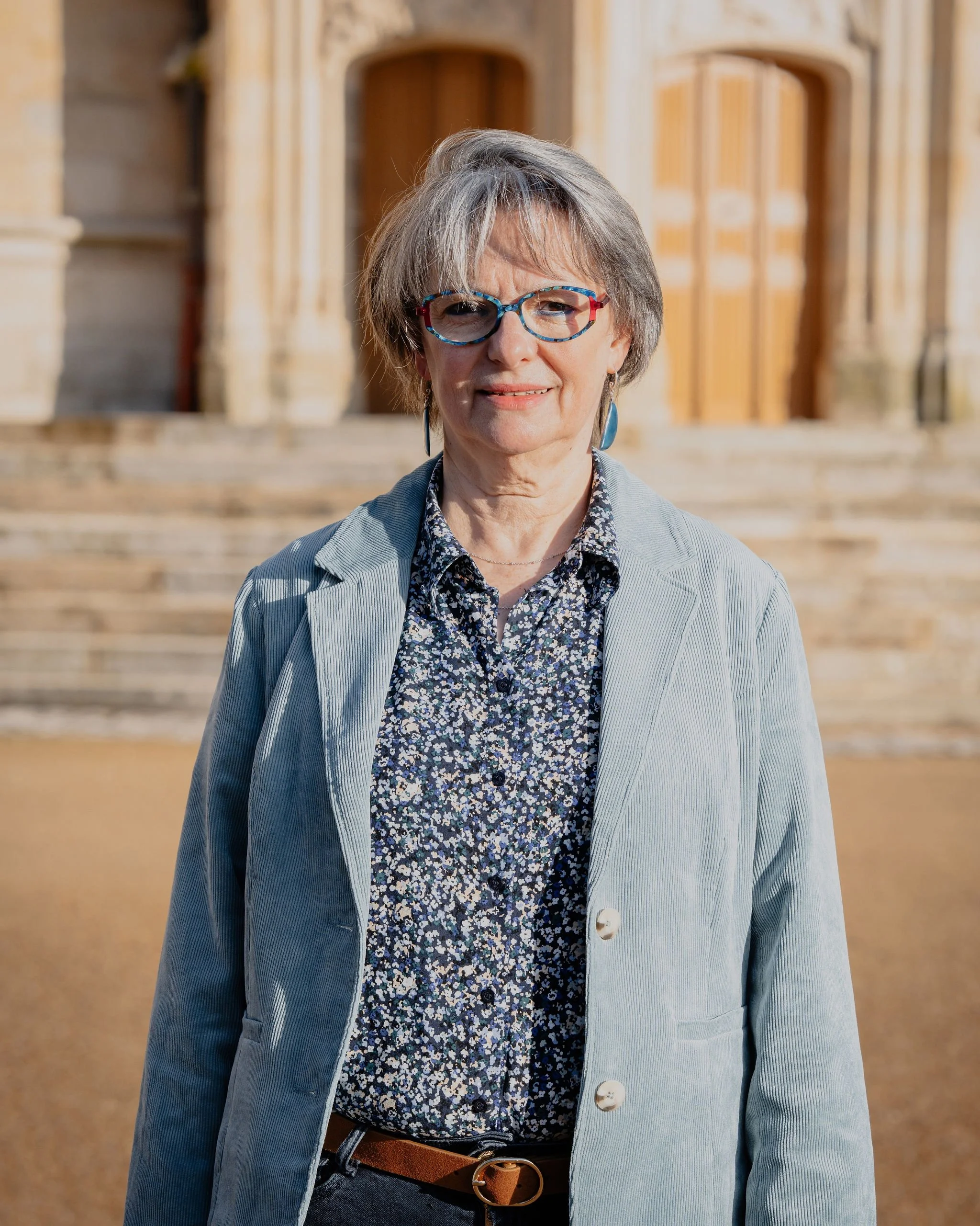 Une femme âgée avec des lunettes colorées, porte une chemise à motifs floraux et un blazer gris, se tient devant une ancien bâtiment en pierre avec des escaliers en marche.